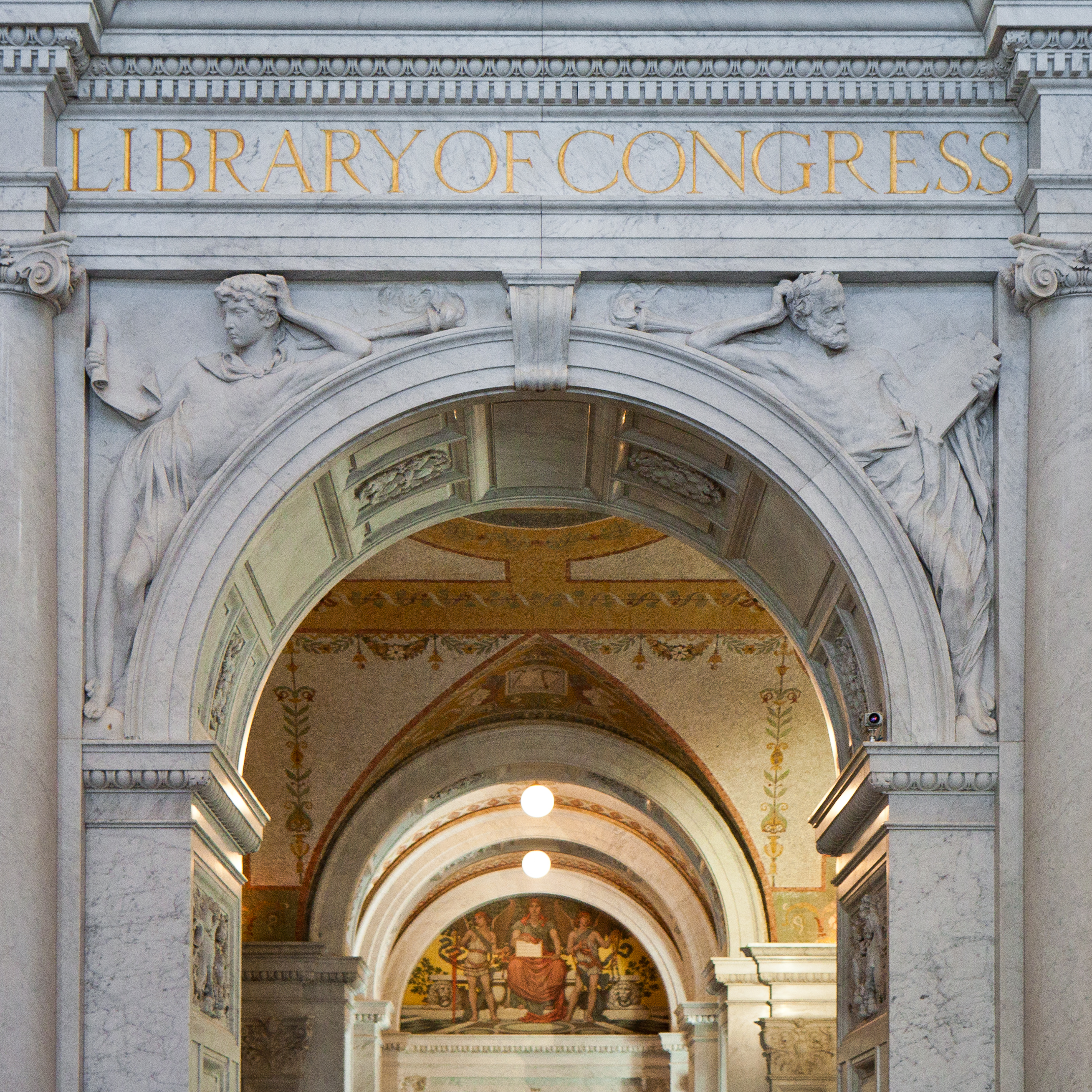  A gray marble arch leading from the Great Hall to the Reading Room at the Library of Congress. In marble on either side of the arch are carvings of two men. On the left is a young student looking at a scroll, on the right is an older man with a beard looking at a stone tablet. Above the arch are the words “Library of Congress.” inscribed in gold.
