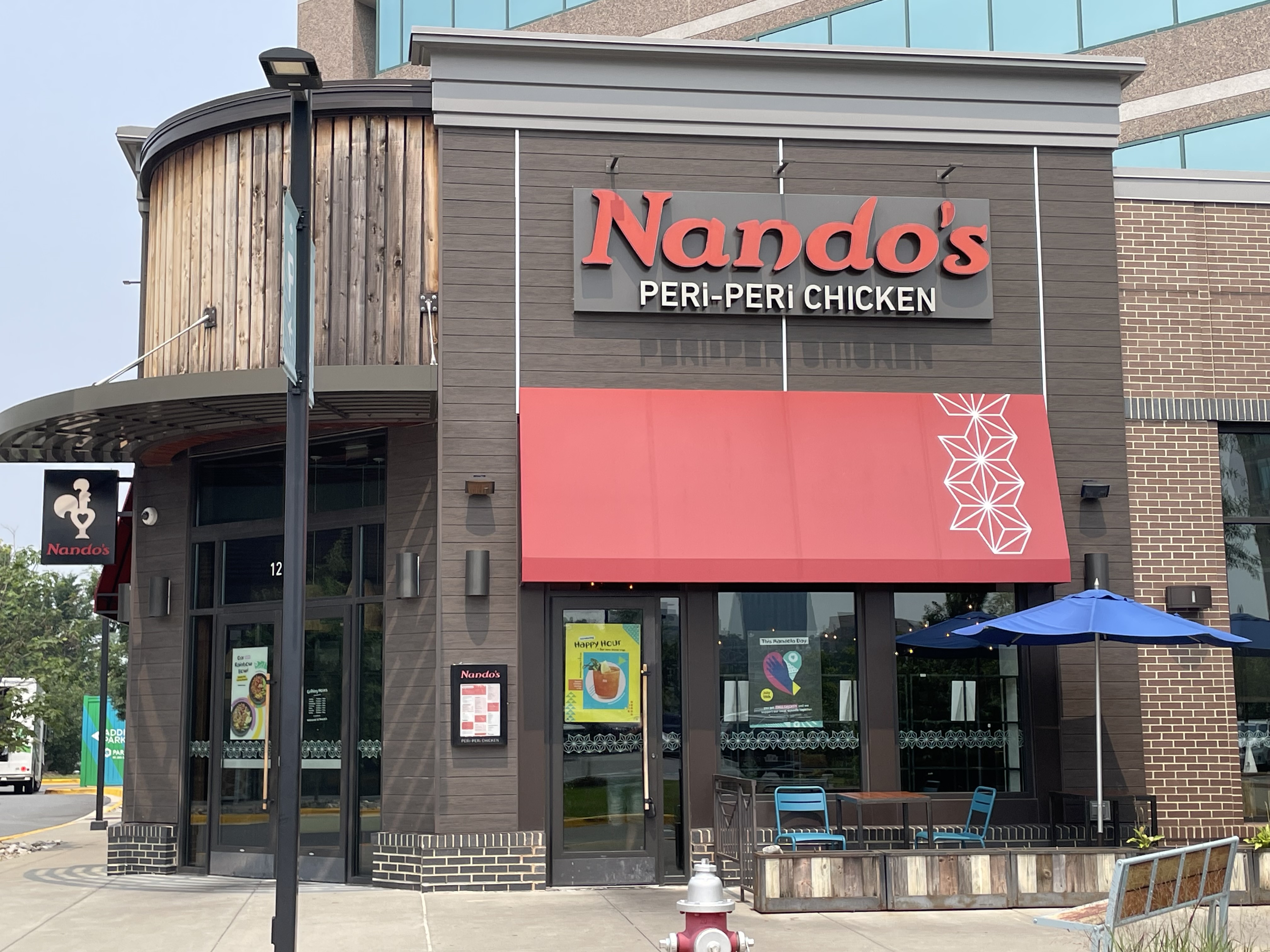Building with brown wooden siding, with “Nando’s Peri-Peri Chicken” sign. Building sits along sidewalk, with small patio and seating in front.