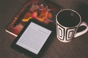 A notebook with glasses and a Kindle on top next to a mug. Photo by Aliis Sinisal via Unsplash