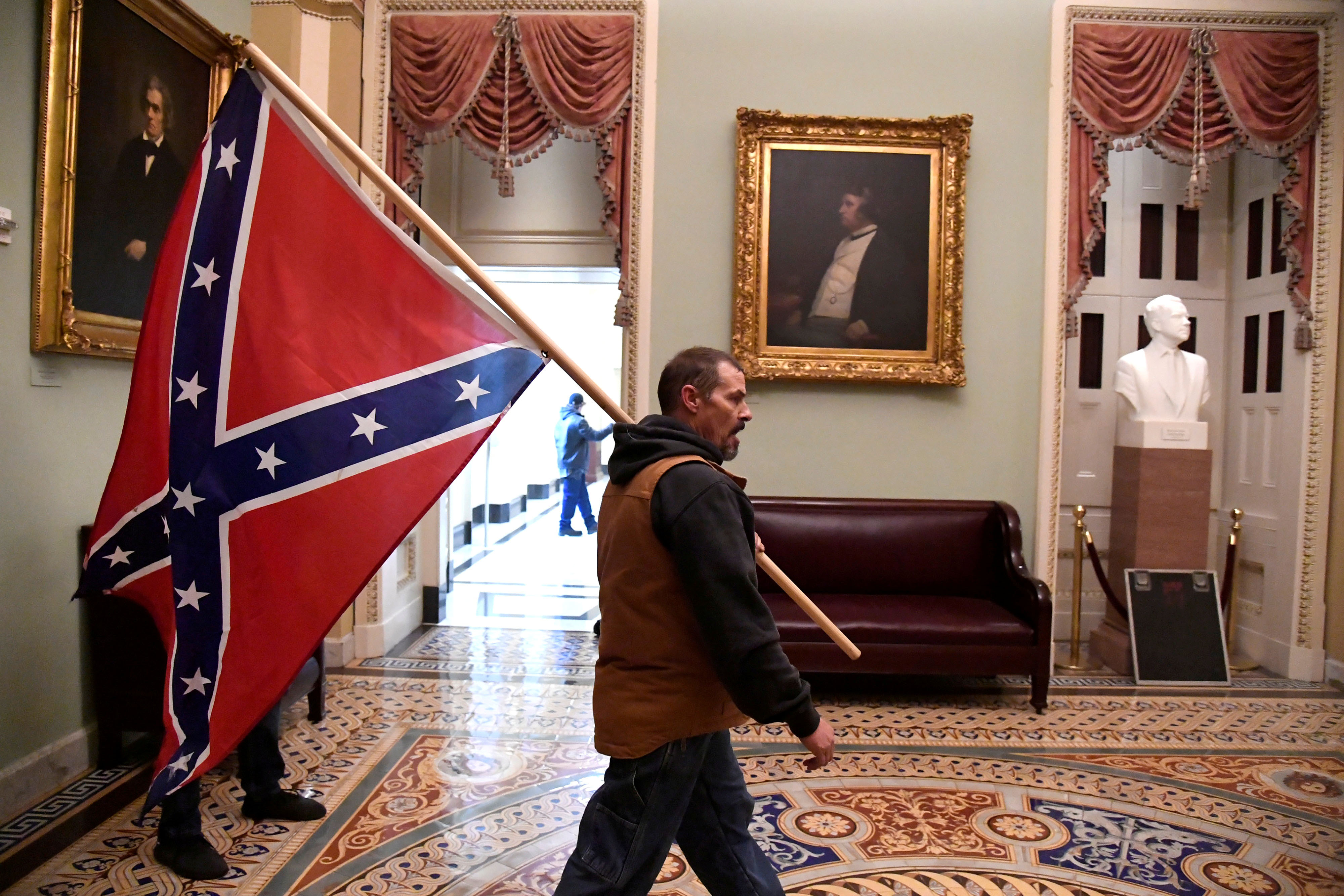 A member of the mob that stormed the US Capitol on January 6 holds a Confederate flag aloft in front of a portrait of John C. Calhoun (left).