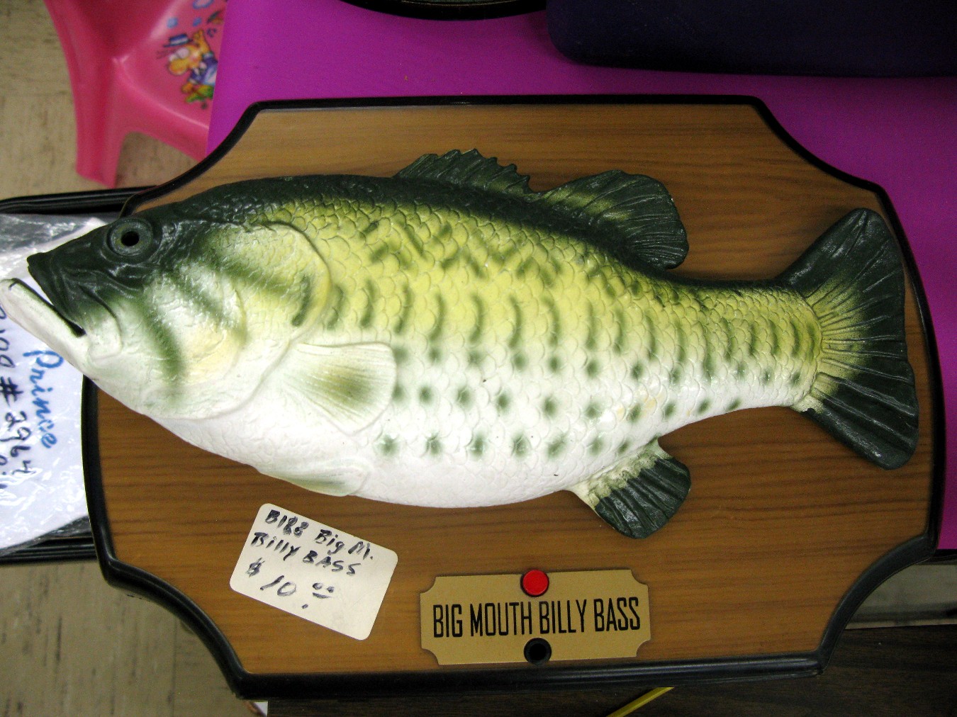 Close-up color photograph of a rubber fish above a red button and a nameplate that says “Big Mouth Billy Bass.” A price tag adorns the plaque the fish is mounted on.