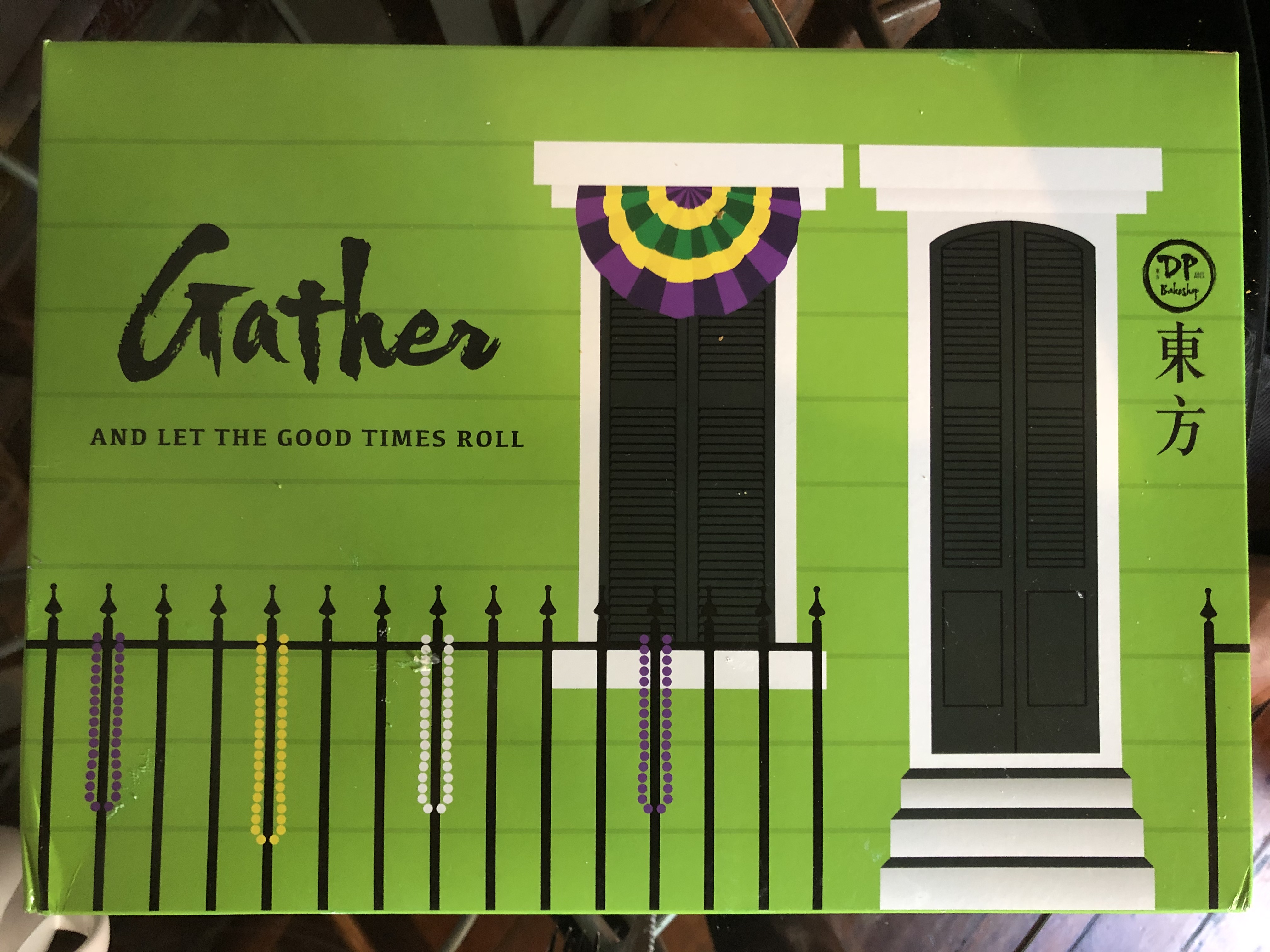 Lime-green box printed with a New Orleans–style house with Mardi Gras beads hanging from an iron fence. Text reads “Gather and let the good times roll” in English. By the house door is a DP Bakery logo and Vietnamese script.