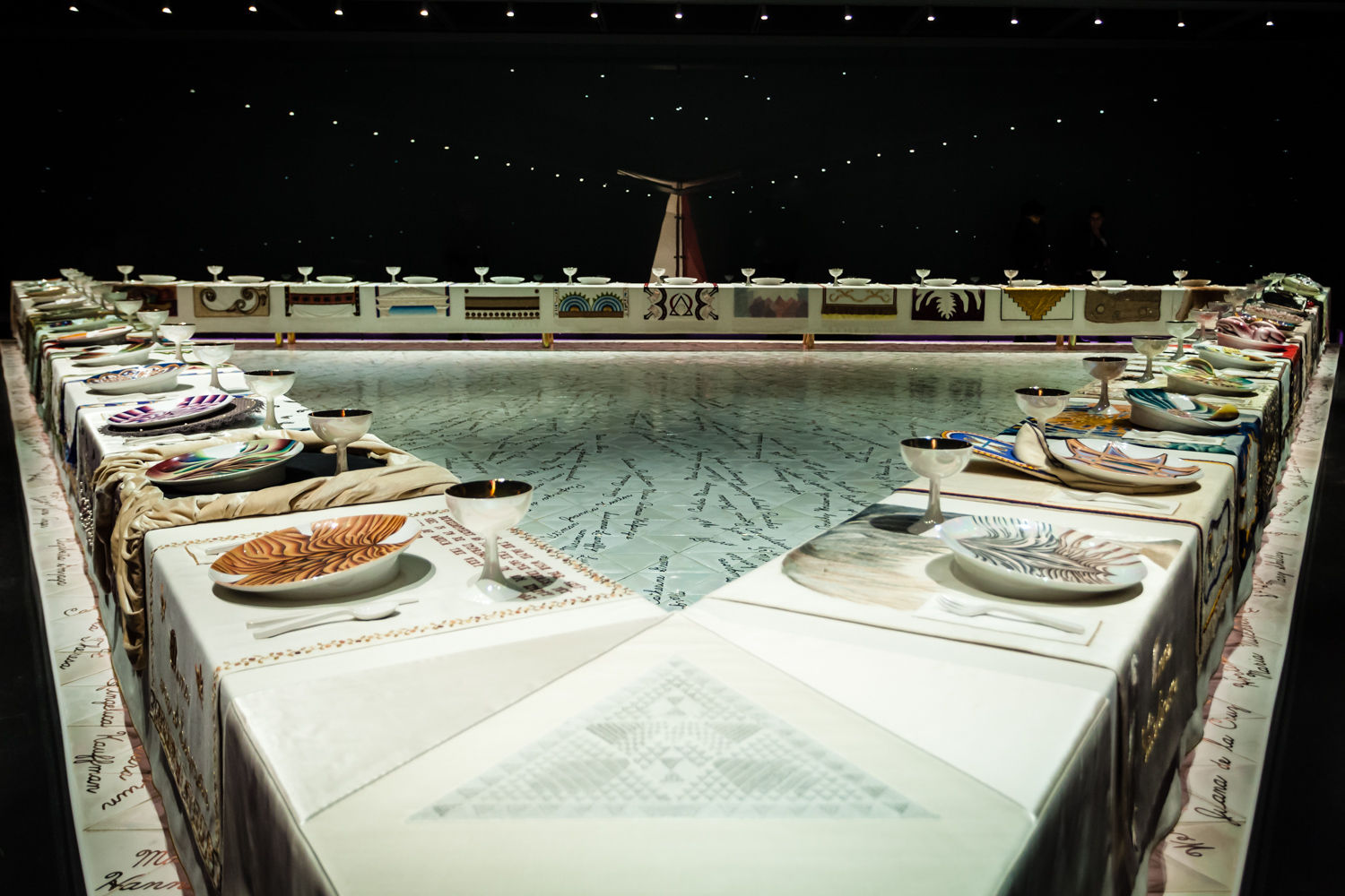 Image of Judy Chicago's art installation The Dinner Party. Depicted is a large banquet table in the shape of a triangle with thirty-nine place settings, embroidered runners, gold chalices and utensils, and china painted porcelain plates. The names of important women from history are inscribed in gold on the white floor below the table.