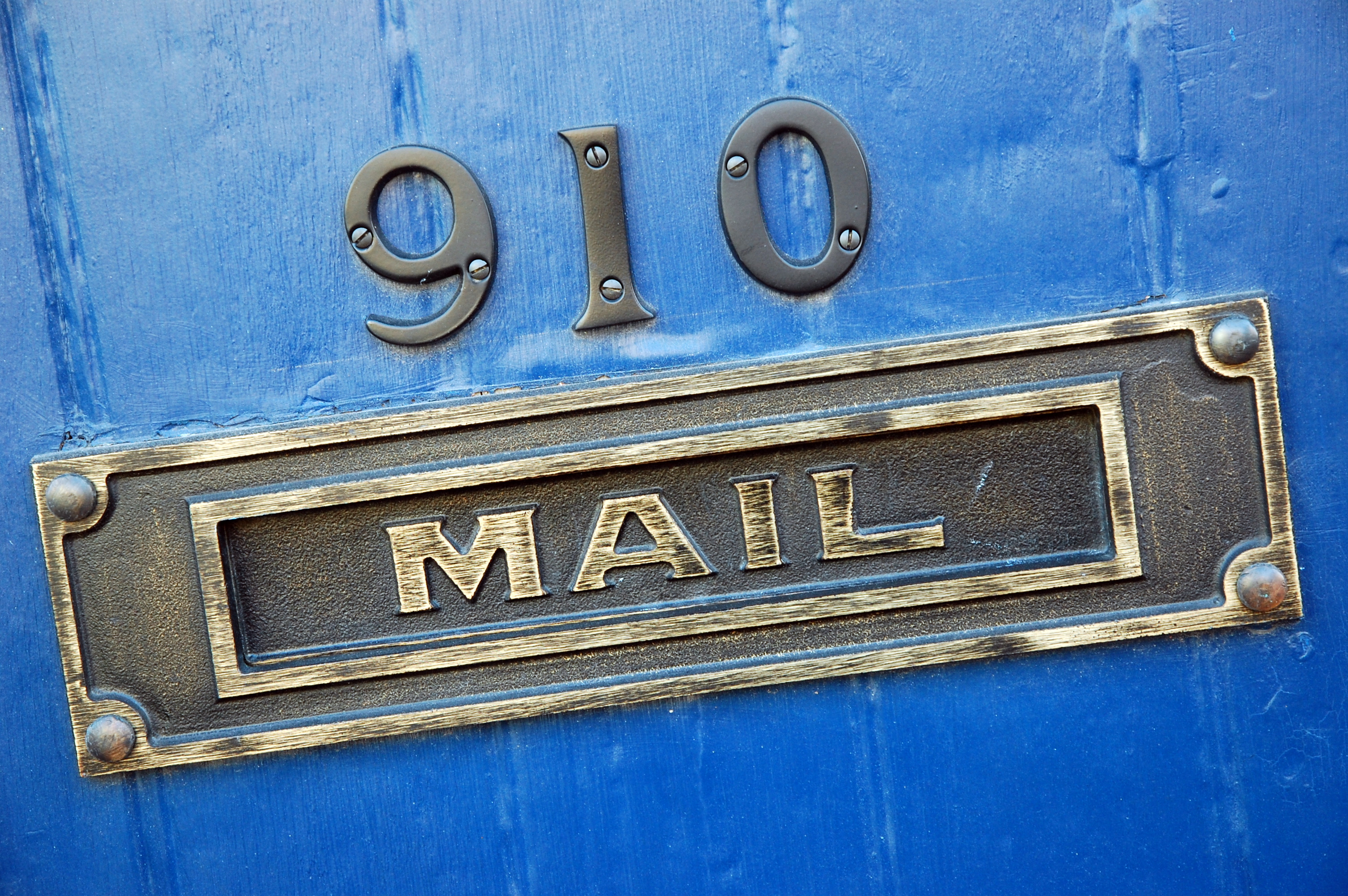 Close-up photograph of a brass mail slot in a door. The door is painted blue and above the mail slot are the numbers 910.