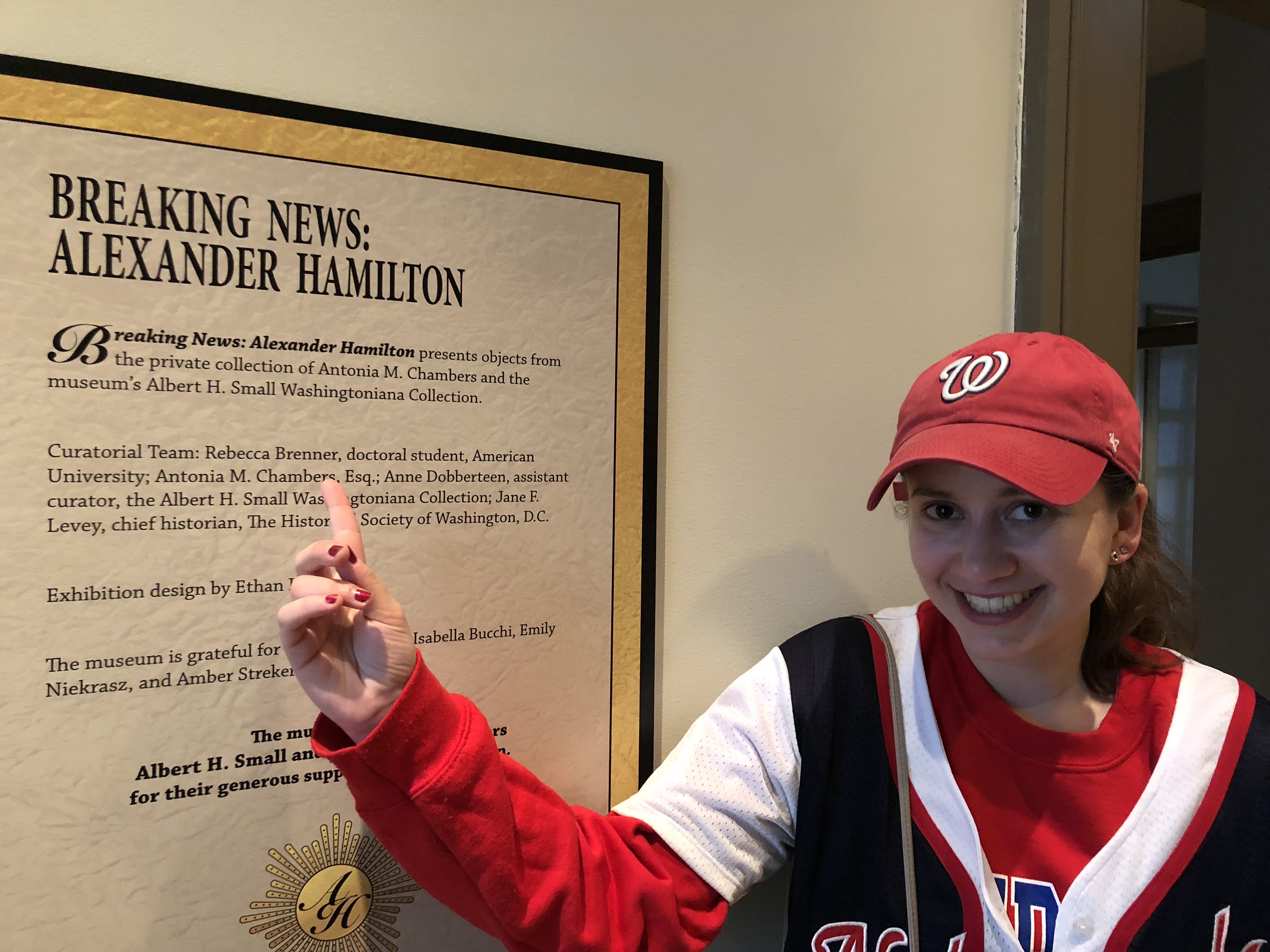 Rebecca Brenner Graham in a red sweatshirt with a white, red, and black jersey on top, and a red ball cap with a white W, the logo for the Washington Nationals.  Brenner is pointing to a poster for “Breaking News: Alexander Hamilton” for the George Washington University Museum. Her finger is pointing to “Curatorial Team: Rebecca Brenner, doctorial student, American University.”