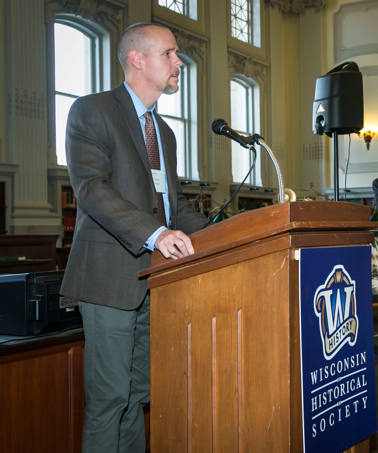 James H. Sweet stands at a lectern with the Wisconsin Historical Society logo on the front in a grey suit, blue button down, and a tie with red and orange squares.