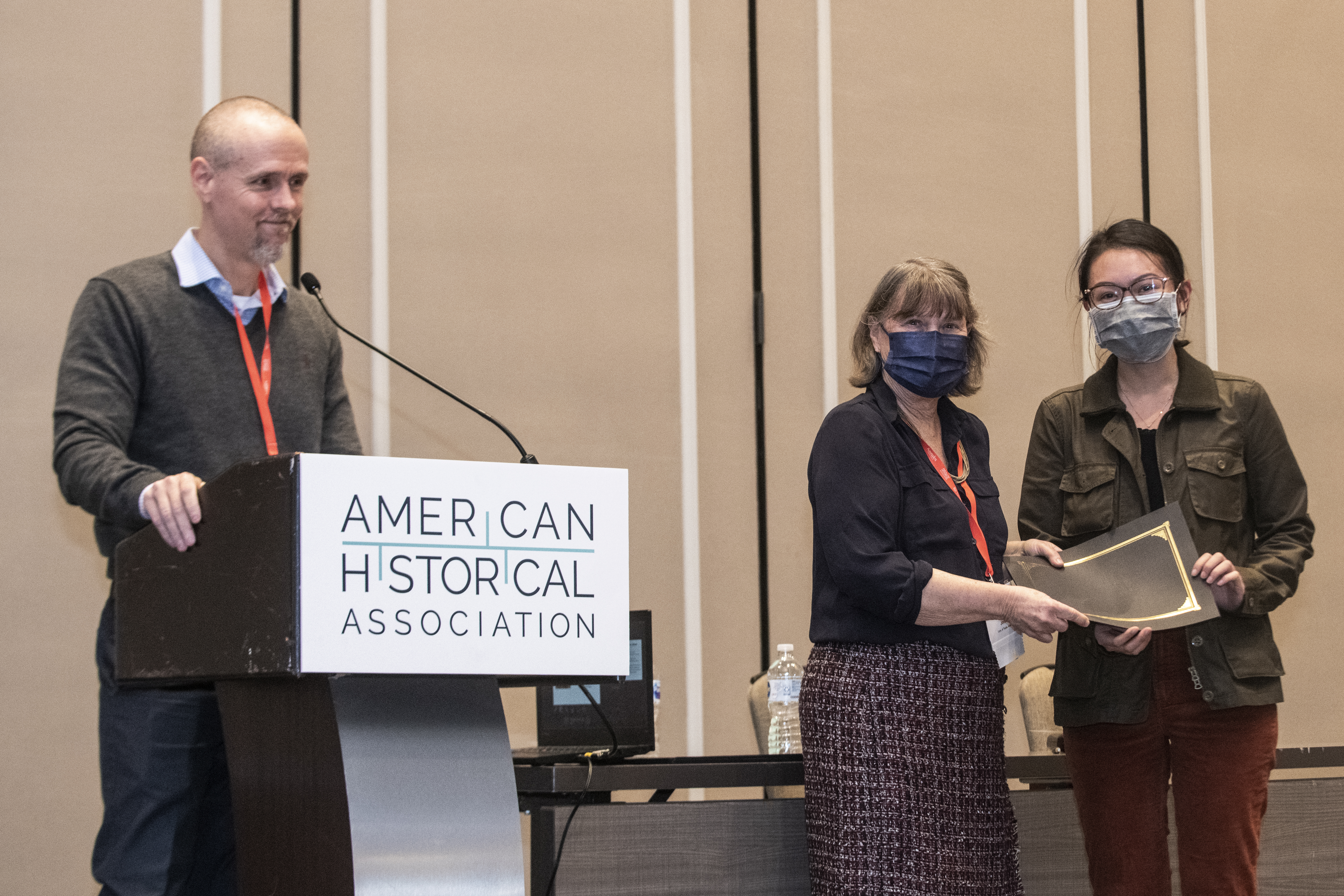 2022 president James H. Sweet stands behind a podium in a sweater, button down and slacks. 2021 president Jacqueline Jones stands next to the podium in a black button down and plaid skirt, and presents the Raymond J. Cunningham Prize to Ann Tran wearing a green jacket and red jeans.