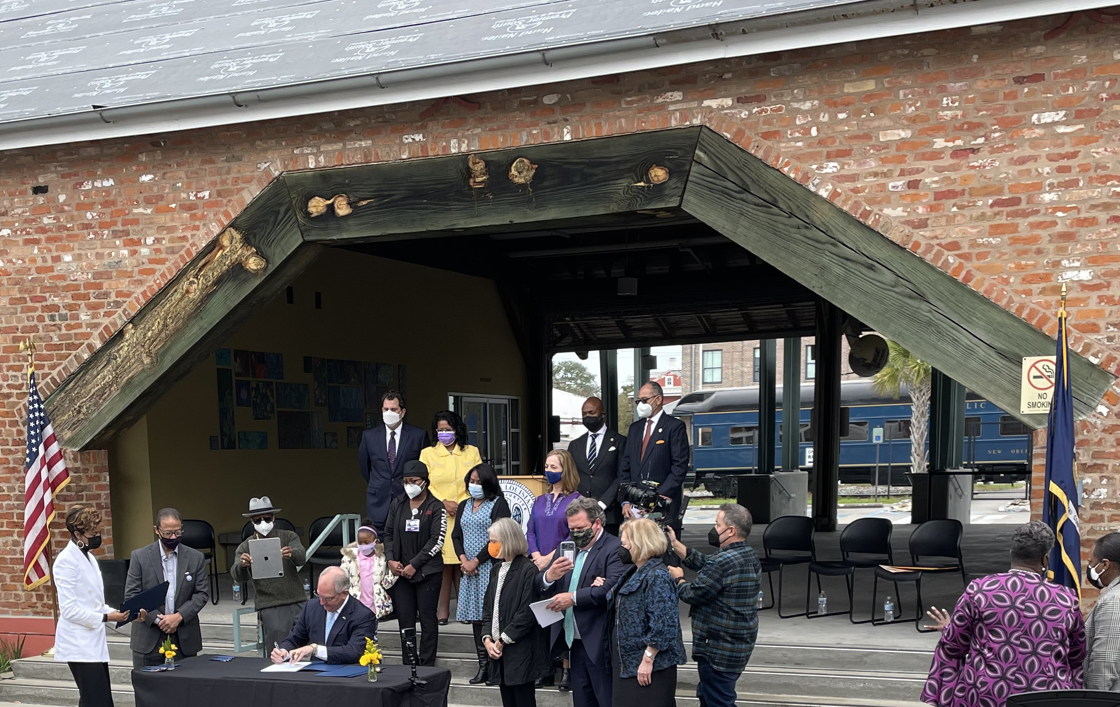 Seated at a table in a black suit and light blue tie, in front of a train, Louisiana Governor John Bel Edwards signs a posthumous pardon for Homer Plessy.He is surrounded by onlookers with face masks standing near and on and outdoor stage with a red brick and wood covering.