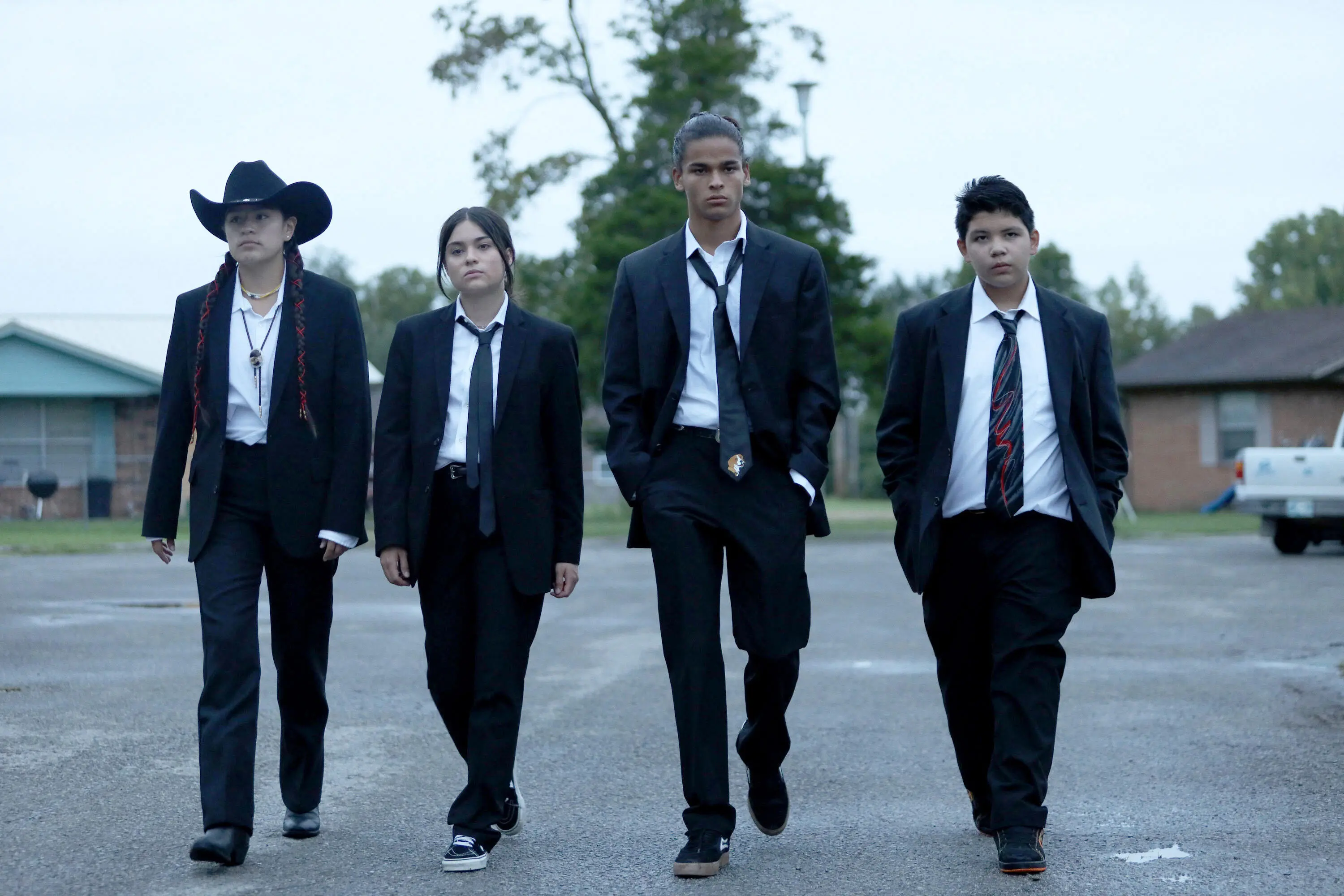 The main characters of Reservation Dogs walk shoulder to shoulder down a street in black and white suits. From left to right Willie Jack, wears a cowboy hat, bolo tie and red string in her two ponytails, Elora wears a black tie and her hair in a ponytail, Bear wears a black tie with an embroidered dog on the end and his hair in a bun, and Cheese wears a black tie with red and white swirls on the front.