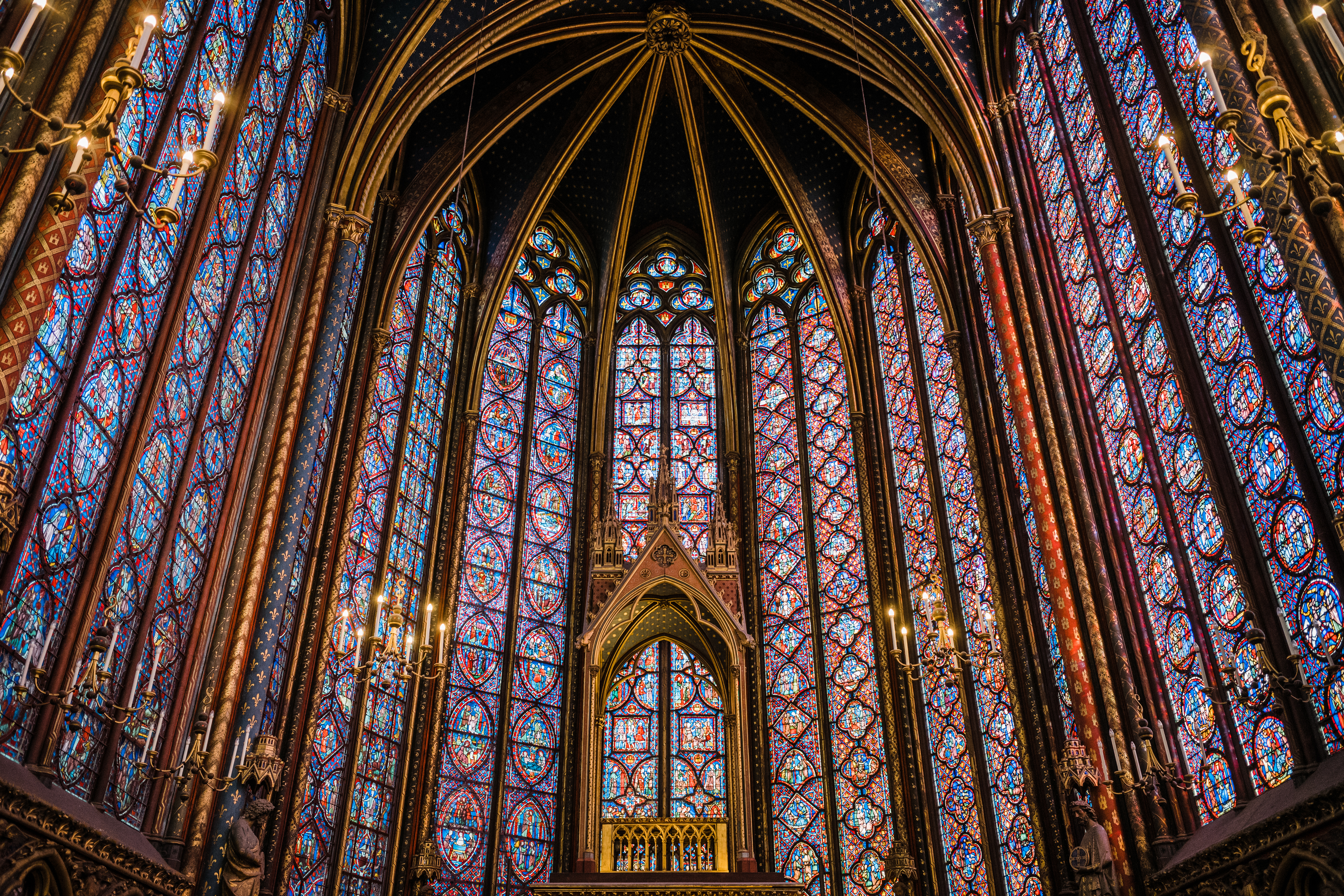 The interior of a chapelle with gold detailing and blue,read,purple, and yellow arched stained glass windows.