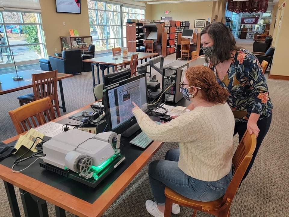  Nancy Locklin-Soferin, a tropical print v-neck shirt and black pants, stands next to her student Laren Gaines. Laren sits at a library table, in a white sweater and light-blue jeans. They are looking at a computer screen, a microfilm machine is on the table next to the computer monitor.
