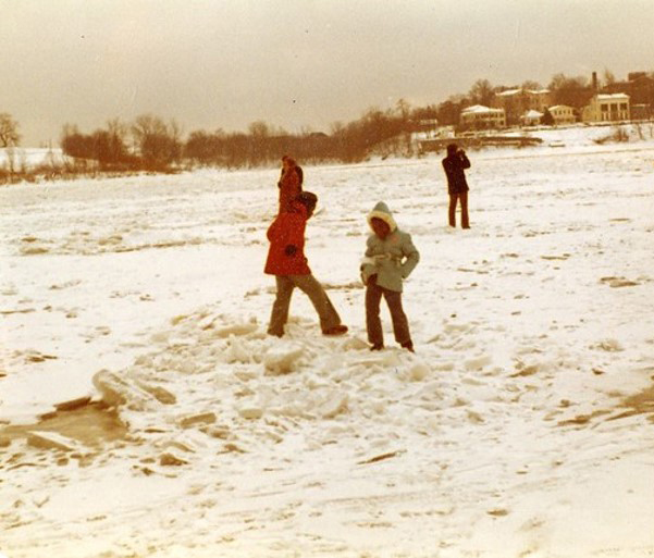 The Ohio River frozen over and covered in white snow, people are walking on the snow and ice. Two kids are playing in the snow, one in a red winter coat, black hat and gloves, the other is in a hooded light blue coat.