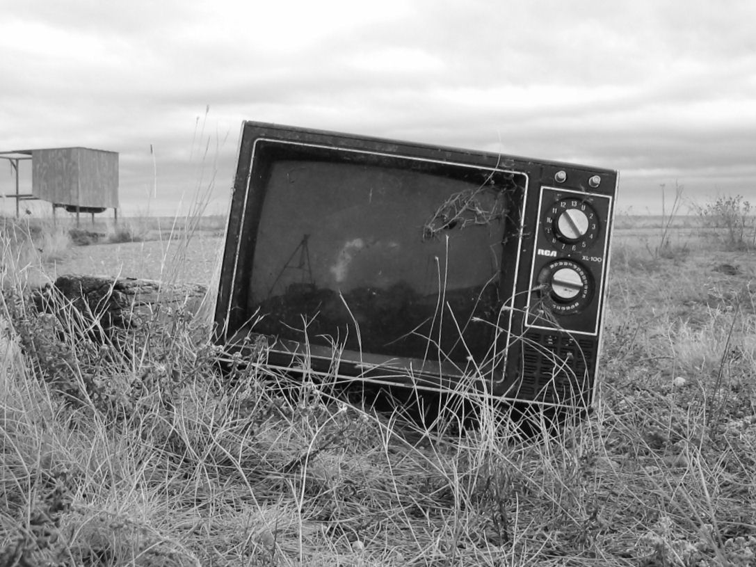 A balck and white image of an old TV with dials, in a field of dead grass. In the background is a wooden shelter.