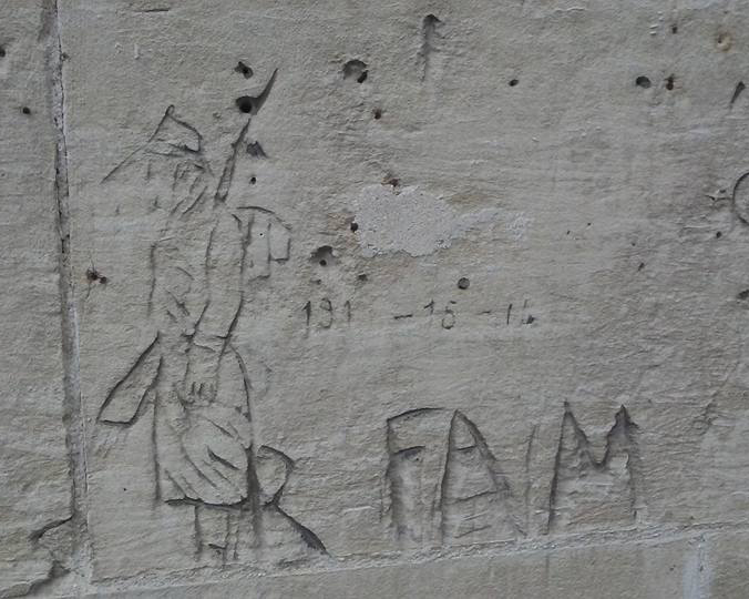 A grey cement wall with a carving of a man in a hat and long coat. Next to the man are the words and the numbers “F. A. M. 1914-15-16” carved into the wall