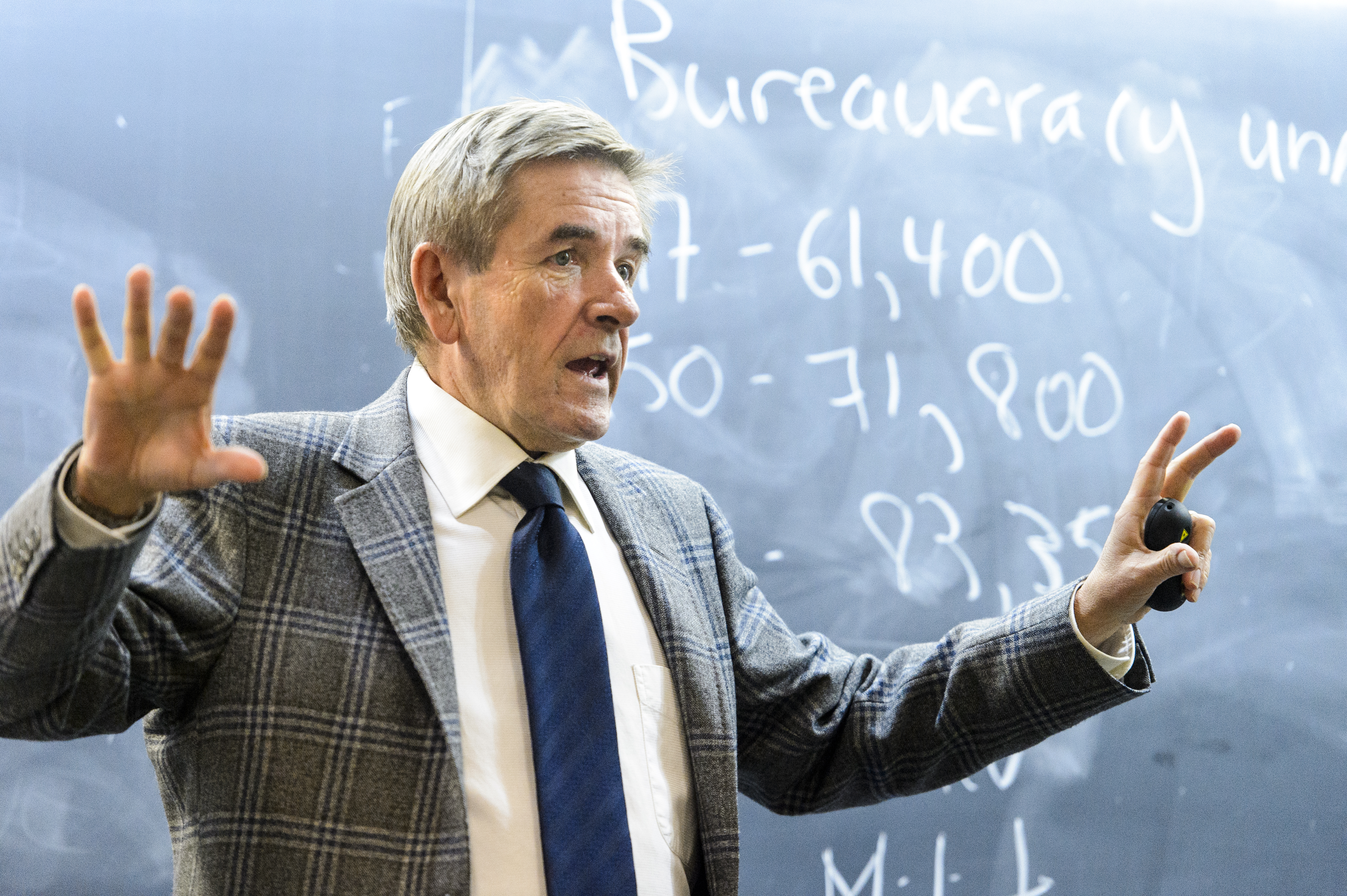 David McDonald, a white man wearing a patterned tweed blazer and blue tie, gestures in front of a chalkboard.