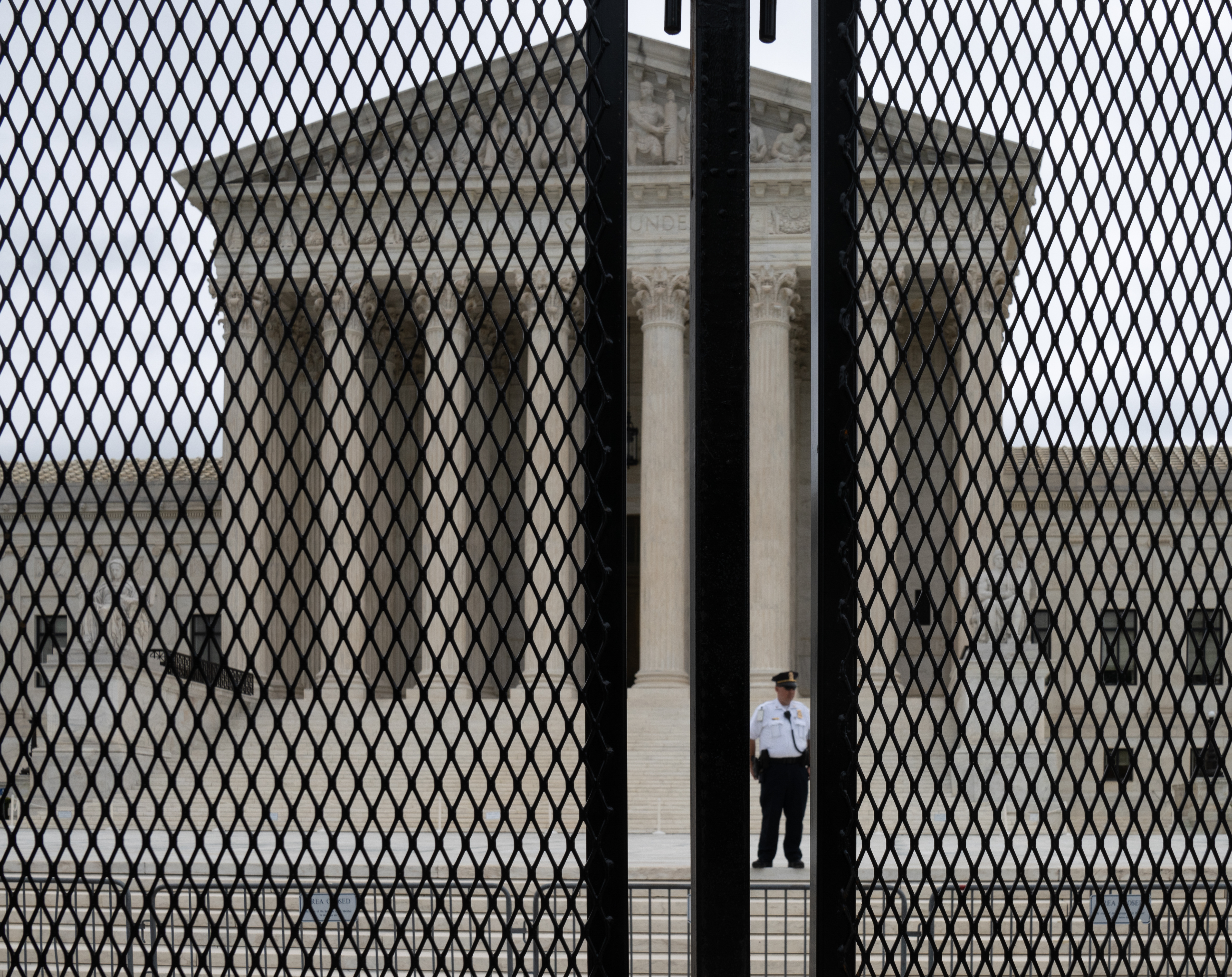 The front of the Supreme Court, a white build with a triangle roof, steps, and columns. A tall black fence sits in front of the building, a security guard is seen through the fence standing at the bottom of the steps to the building.