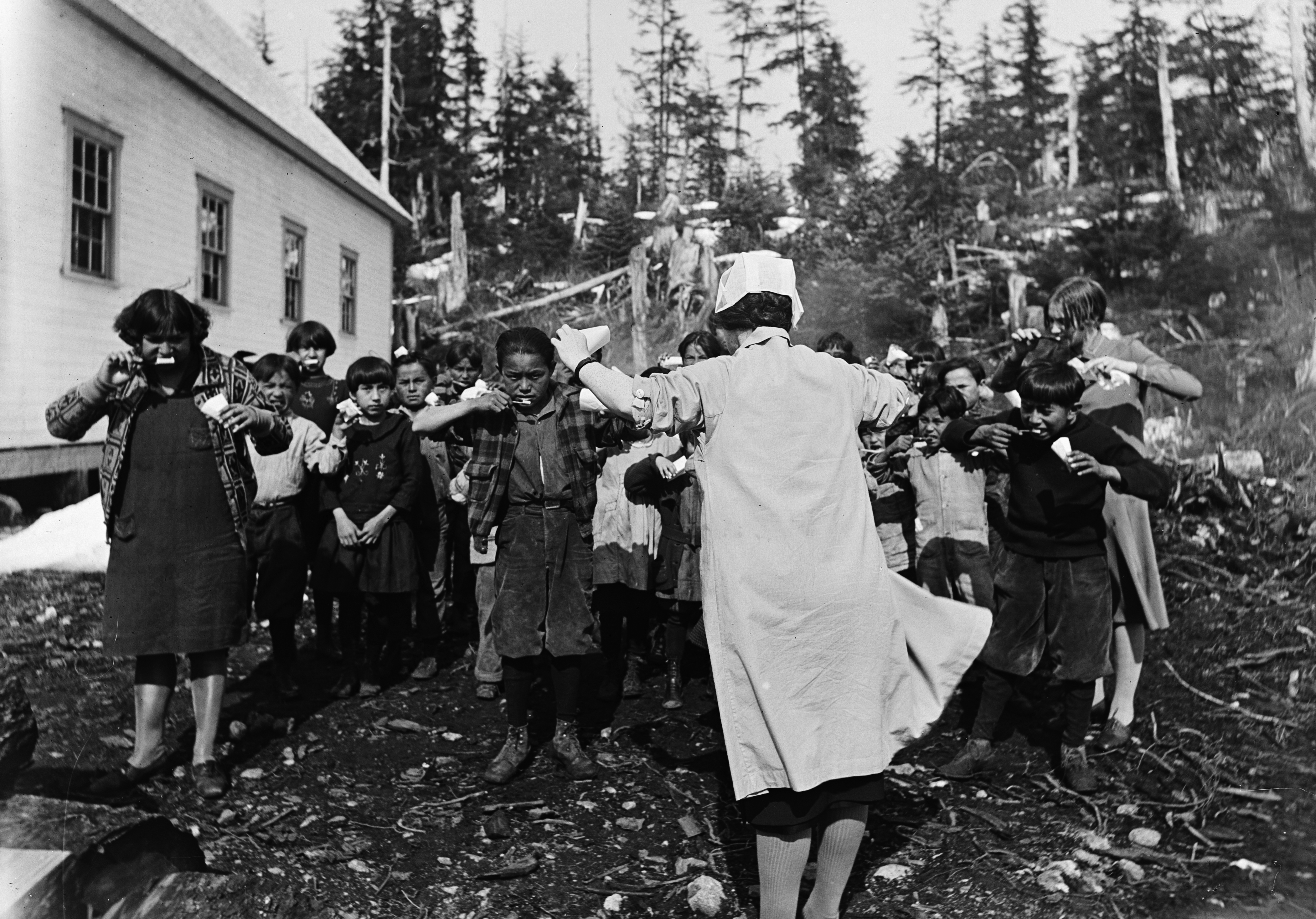 Black and white photograph of a white nurse teaching a group of Alaska Native children how to brush their teeth outside of a Mission School.