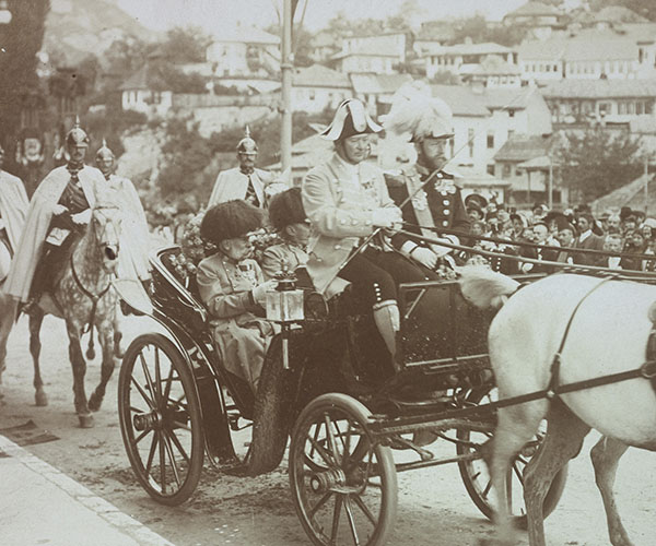 An elderly man with impressive facial hair, dressed in a military uniform and flanked by mounted soldiers, parades by a watching crowd in a black four-wheeled carriage pulled by two white horses. It is captioned “Kaiser Franz Josef I. in Sarajevo” in black letter print.