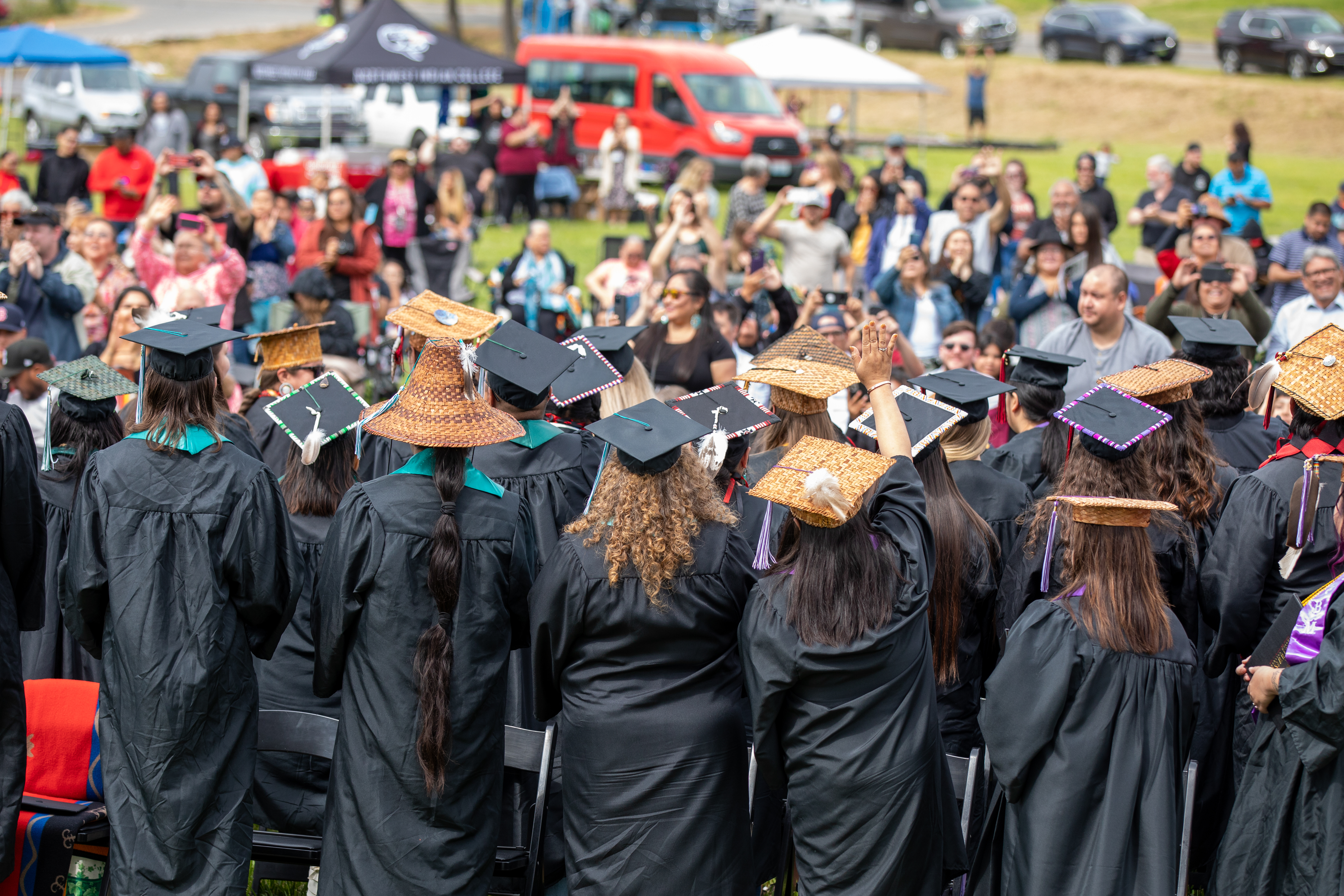 The backside of a group of students at their graduation wearing regalia. They are waving and looking at adults taking pictures of them.