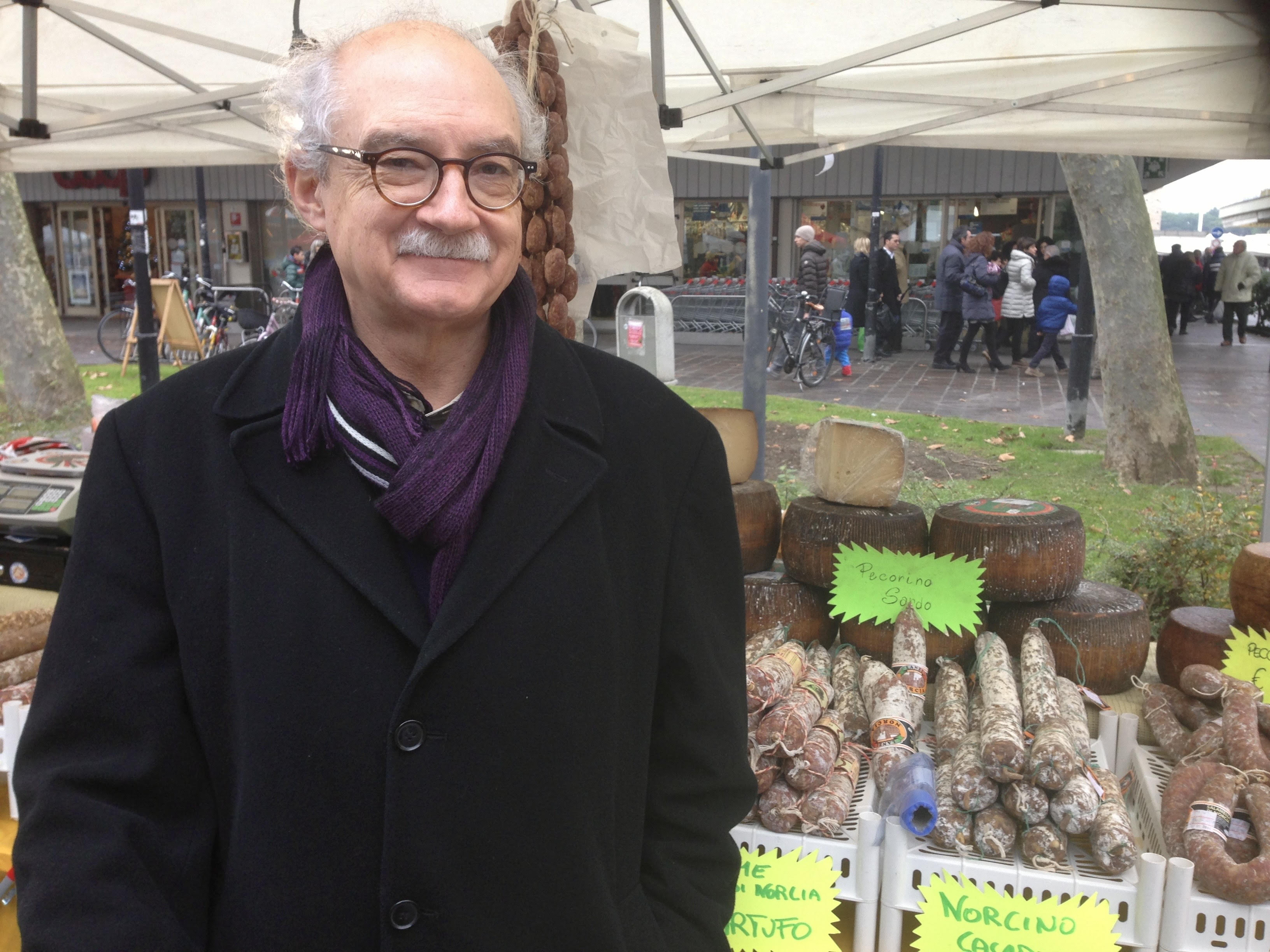 A white man with gray hair, glasses, and a mustache stands in front of a row of cured meats and cheeses at an outdoor market. He wears a black wool coat with a purple scarf.