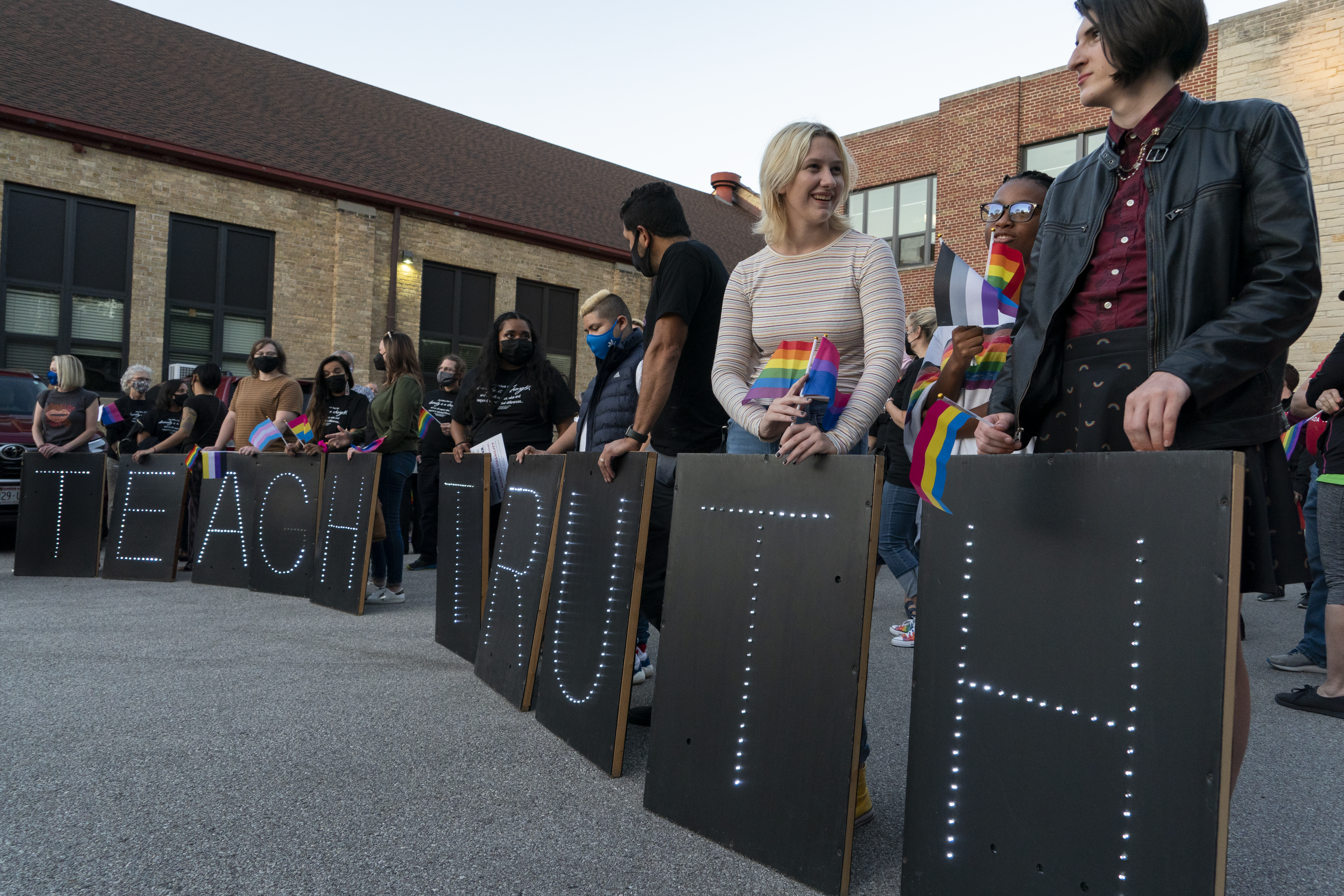 A group of people standing in a line wearing rainbow masks and holding signs in front of them that spell out 'Teach Truth'