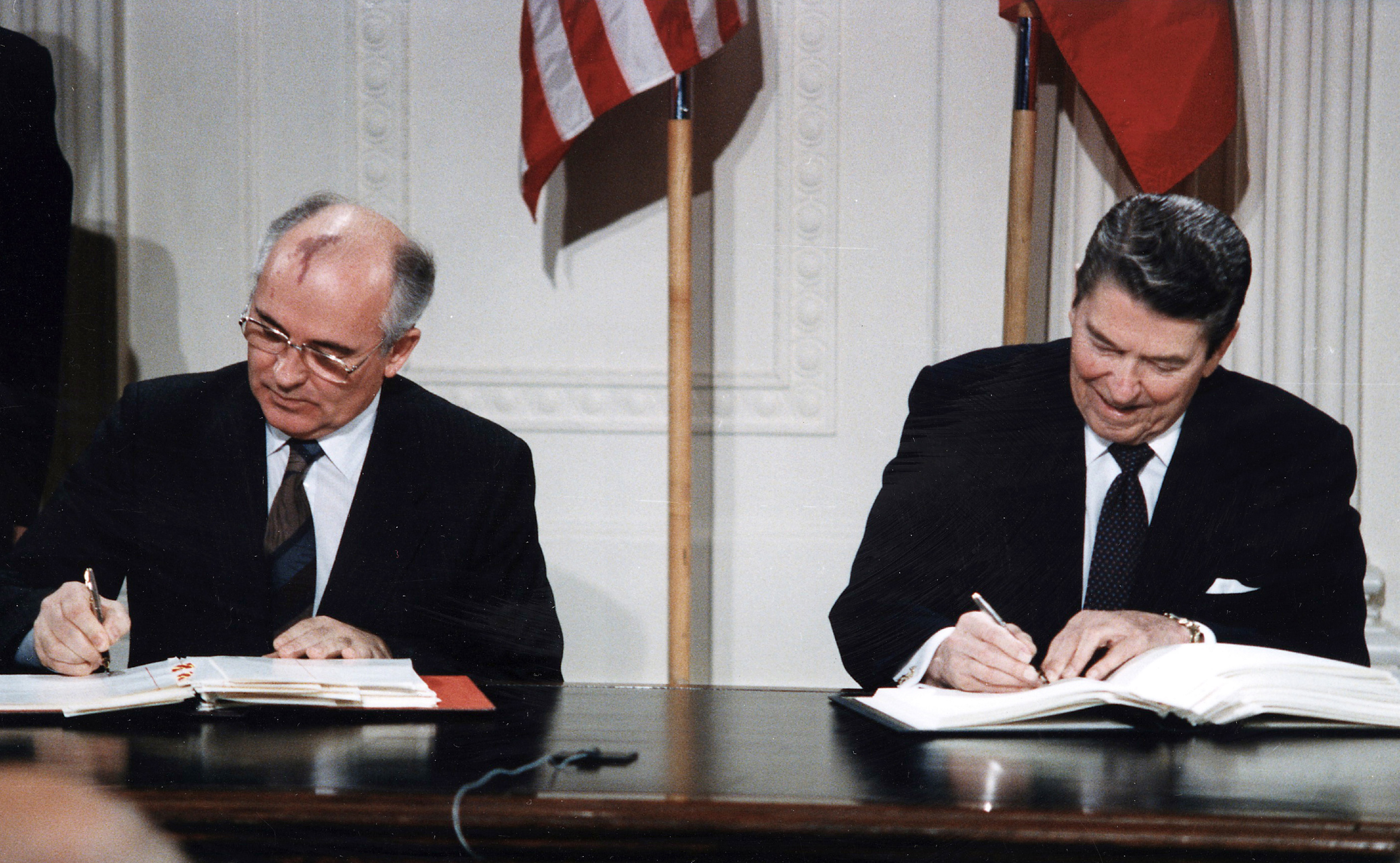 Mikhail Gorbachev and Ronald Reagan sit behind a dark wooden desk in the East Room of the White House, signing the Intermediate-Range Nuclear Forces Treaty.