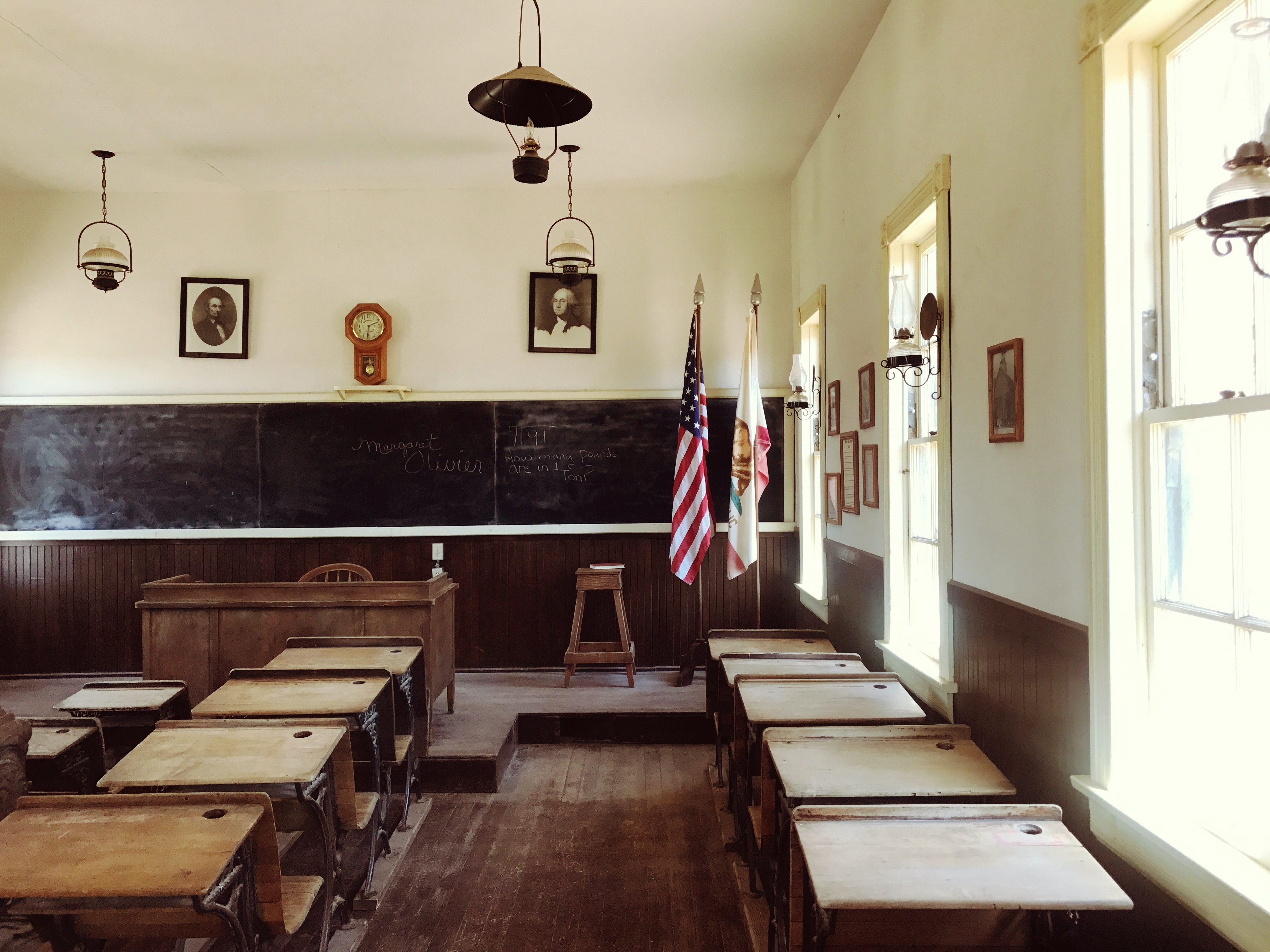 An empty classroom showing rows of desks facing a chalkboard, a teachers' desk, and 2 flags