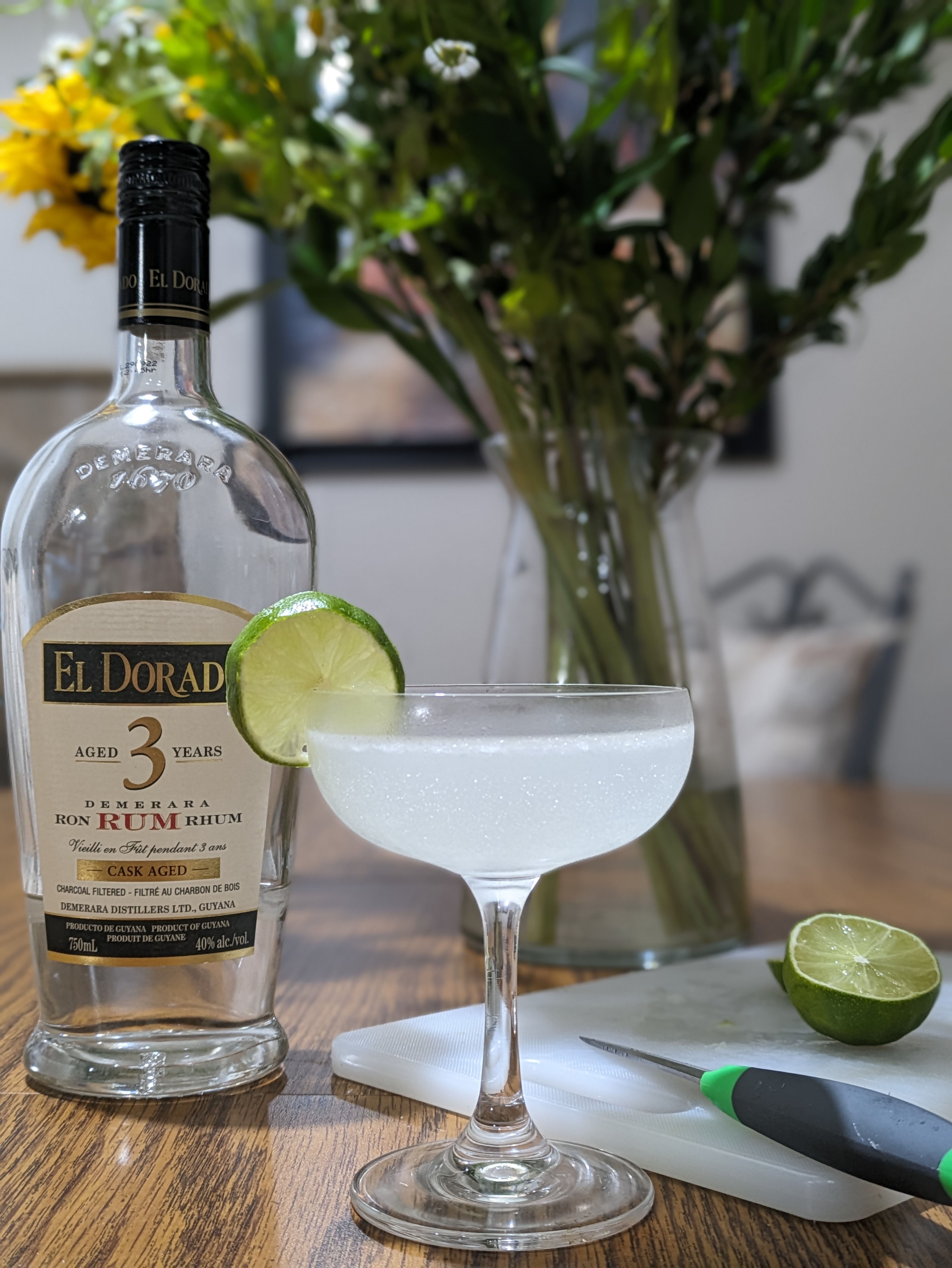 A bottle of El Dorado rum and a cutting board with half a lime stand behind a glass containing a pale liquid and garnished with a lime wheel.