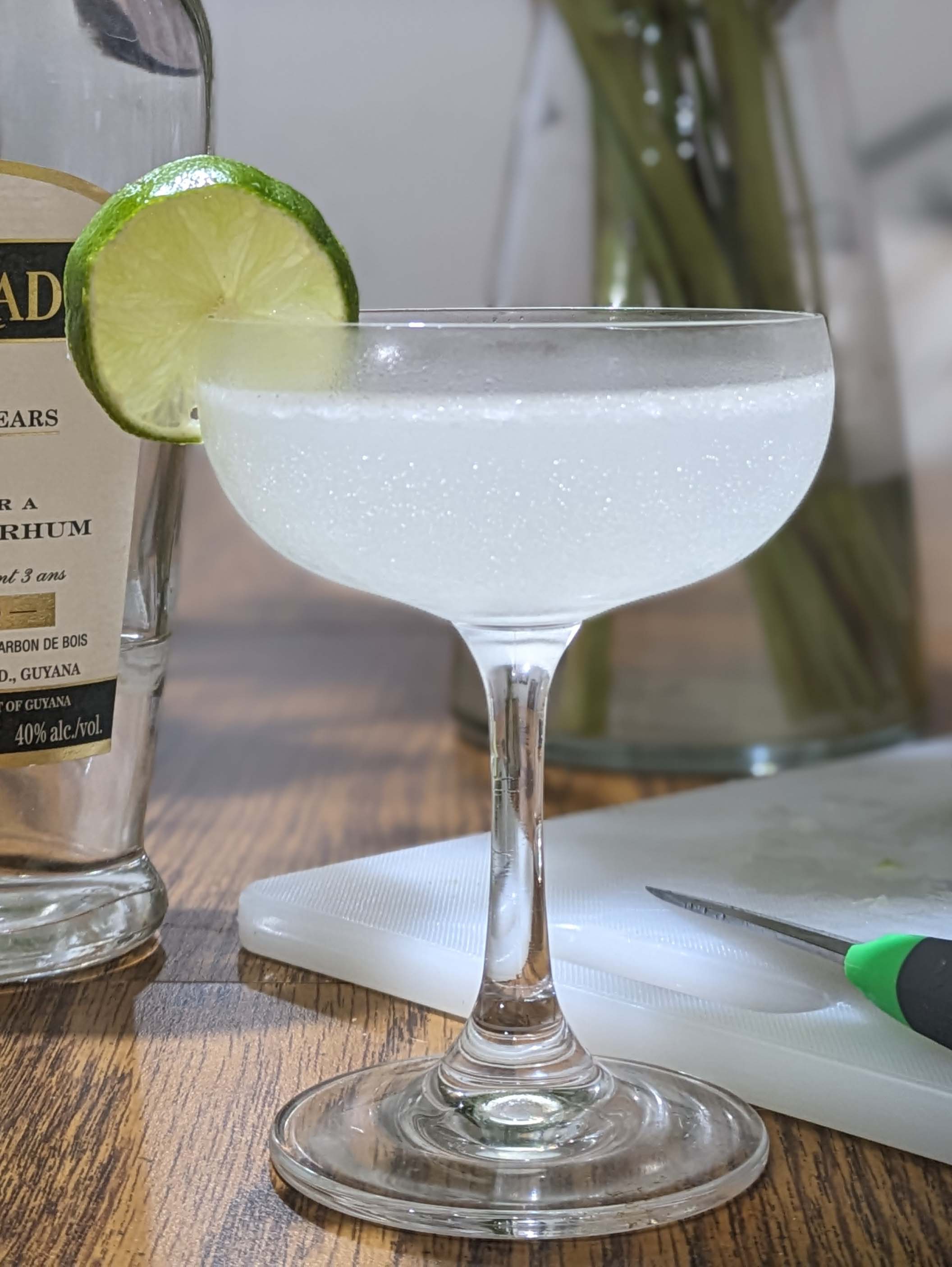 A bottle of El Dorado rum and a cutting board with half a lime stand behind a glass containing a light yellow liquid and garnished with a lime wheel.