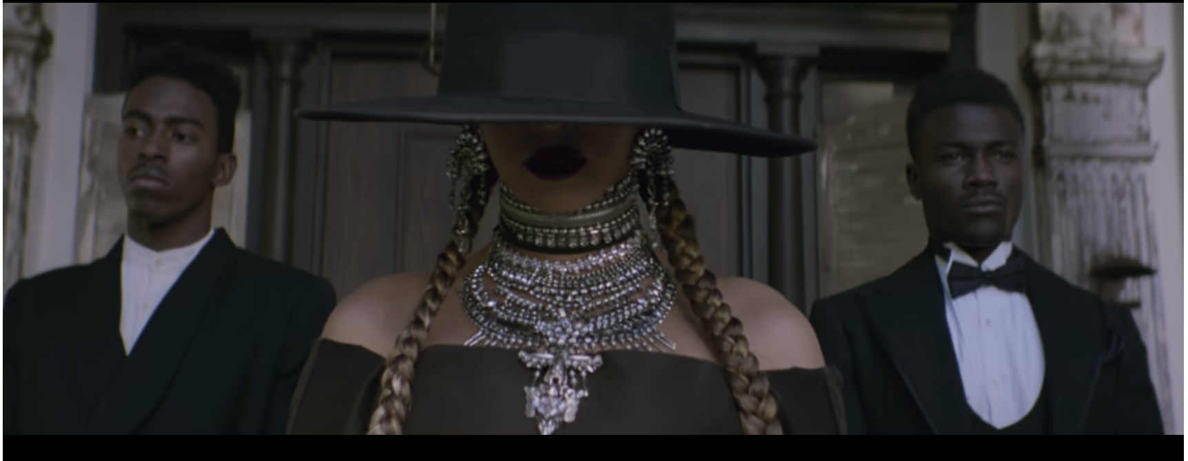 A Black woman wearing a black hat that covers the top half of her face, dark purple lipstick, her hair in two braids, and many silver chunky necklaces stacked from the top of her neck to her chest. She is standing between two Black men staring ahead.