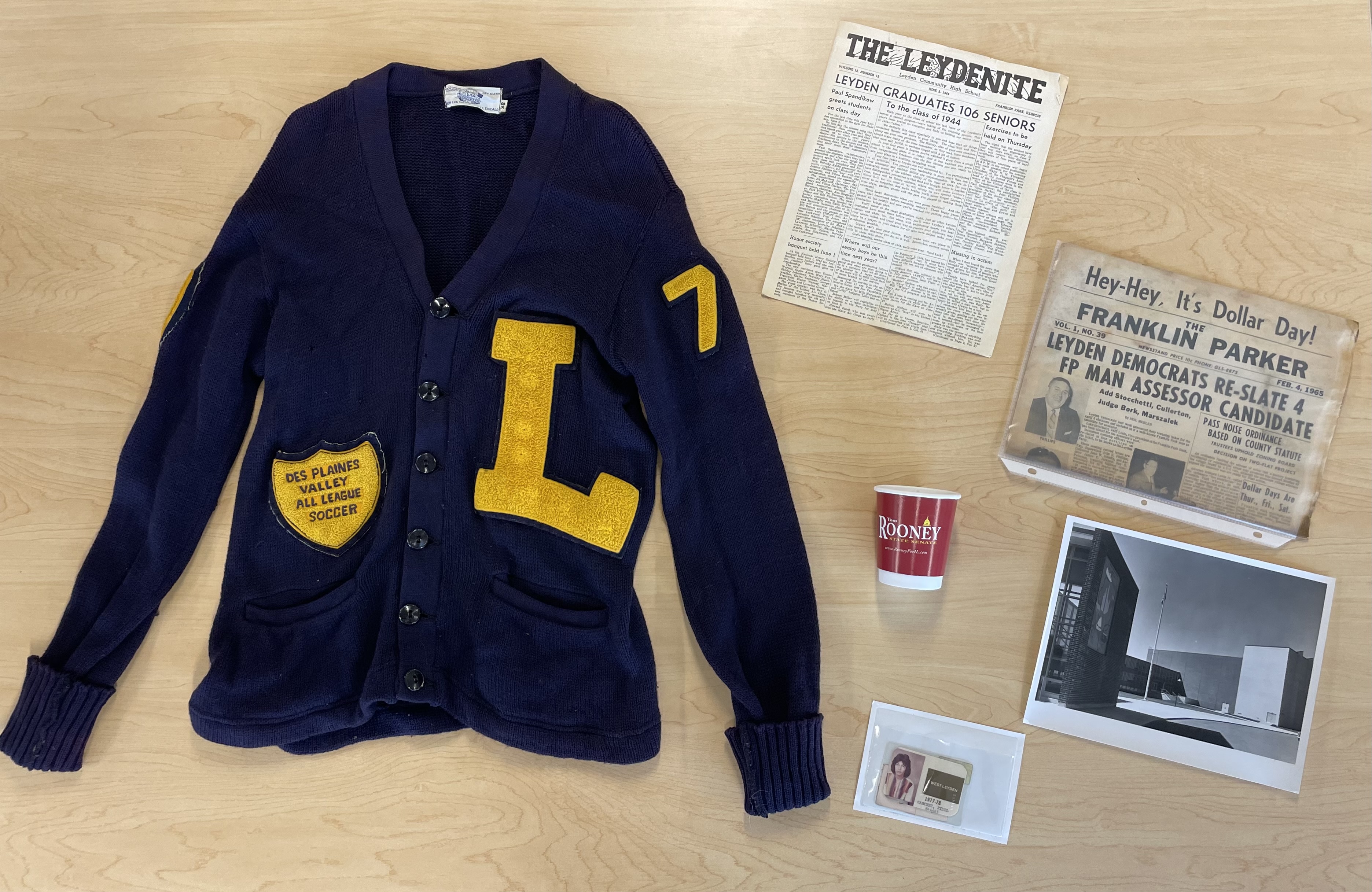 A letterman jersey laid out on a table next to two local newspapers from 1944 and 1965, a paper cup labeled 'Tim Rooney State Senate,' a photo of a building, and a student ID.