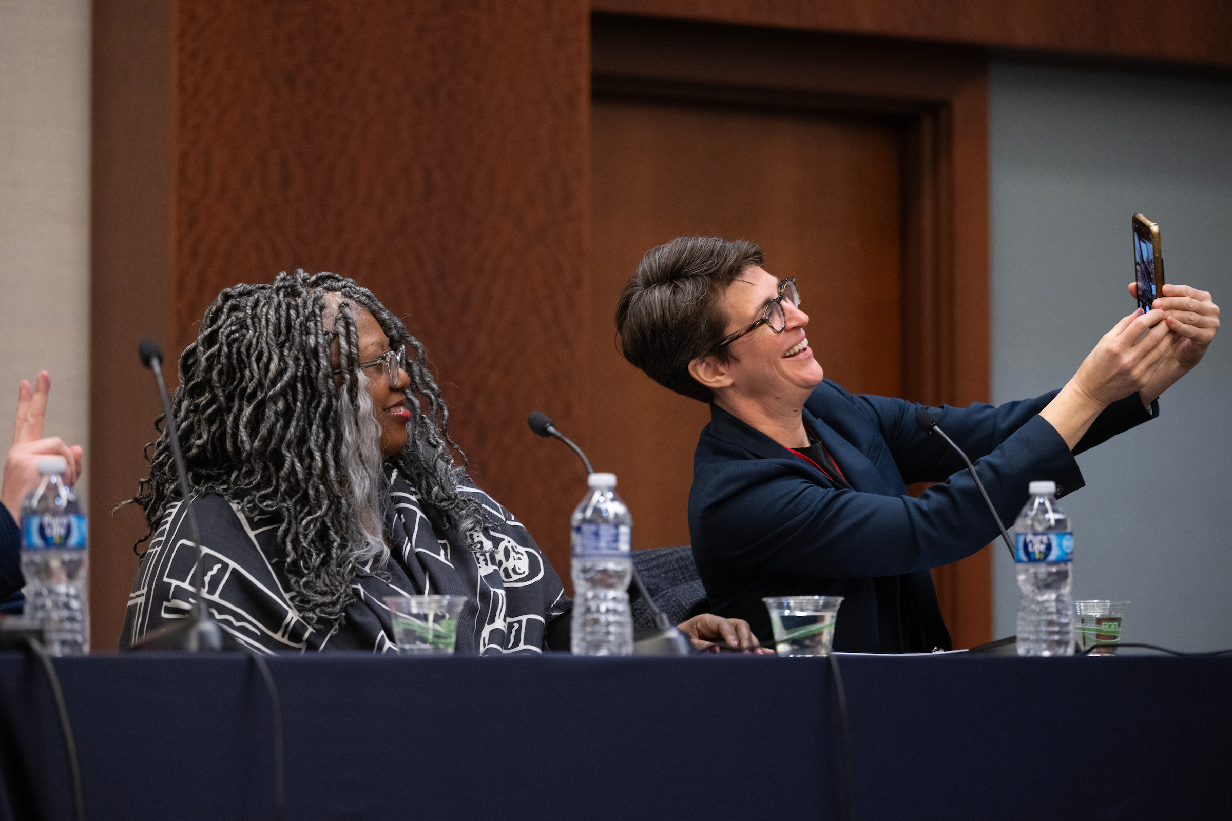 Two women at a panel with the one on the right holding up her cellphone to take a selfie with the other woman