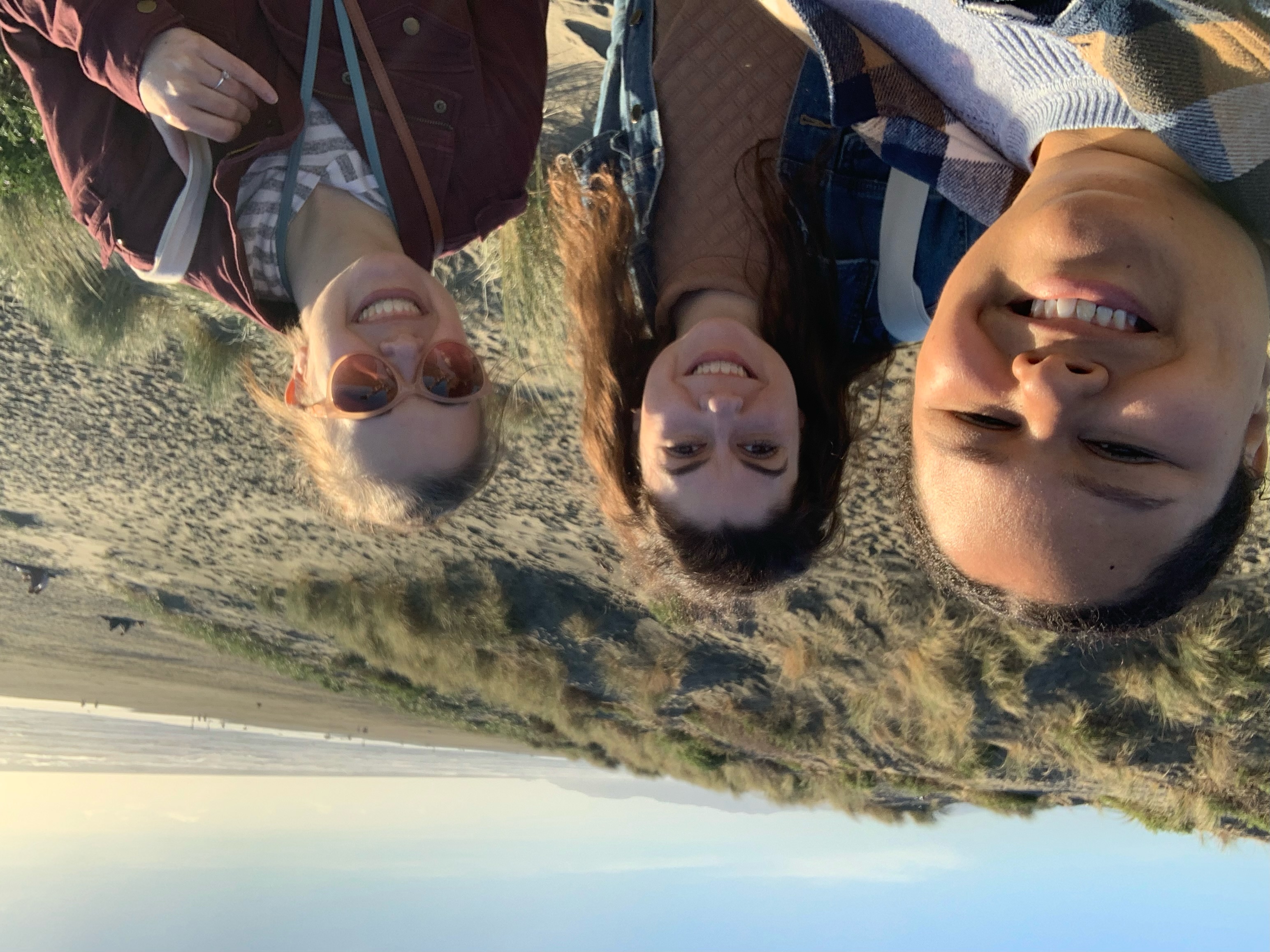 A selfie of three women smiling with a beach behind them
