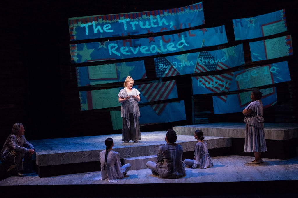 picture of a play with five people on a stage watching one person standing at the center of the stage. There is a painted background behind them that says "the truth revealed" over two American flags