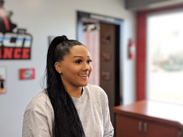 A woman with long black hair and wearing a gray sweatshirt smiles to someone off camera.