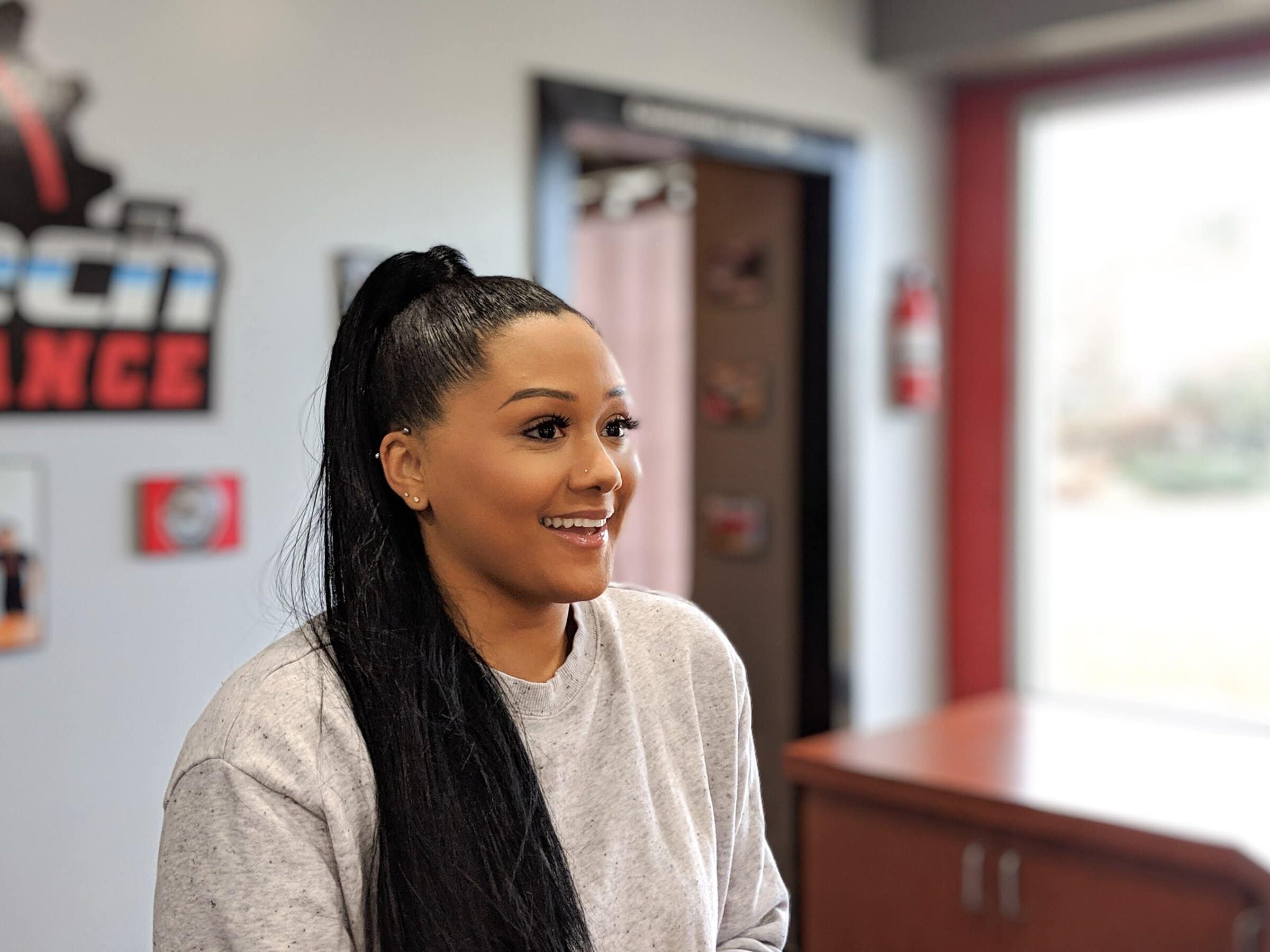 A woman with long black hair and wearing a gray sweatshirt smiles to someone off camera.