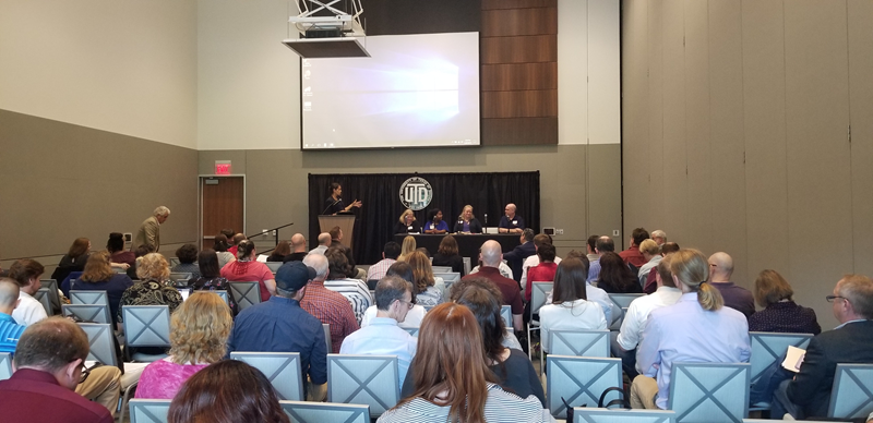 Conference attendees sit in the audience facing a stage with four panelists sitting behind a table with microphones, wearing black and purple shirts. A moderator stands behind a wood podium and faces the panelists to speak to them.