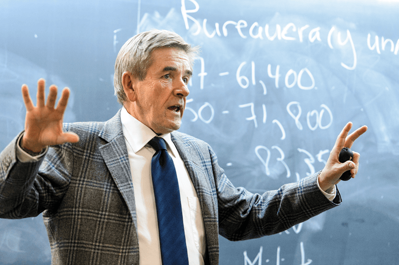 David McDonald, a white man wearing a patterned tweed blazer and blue tie, gestures in front of a chalkboard.