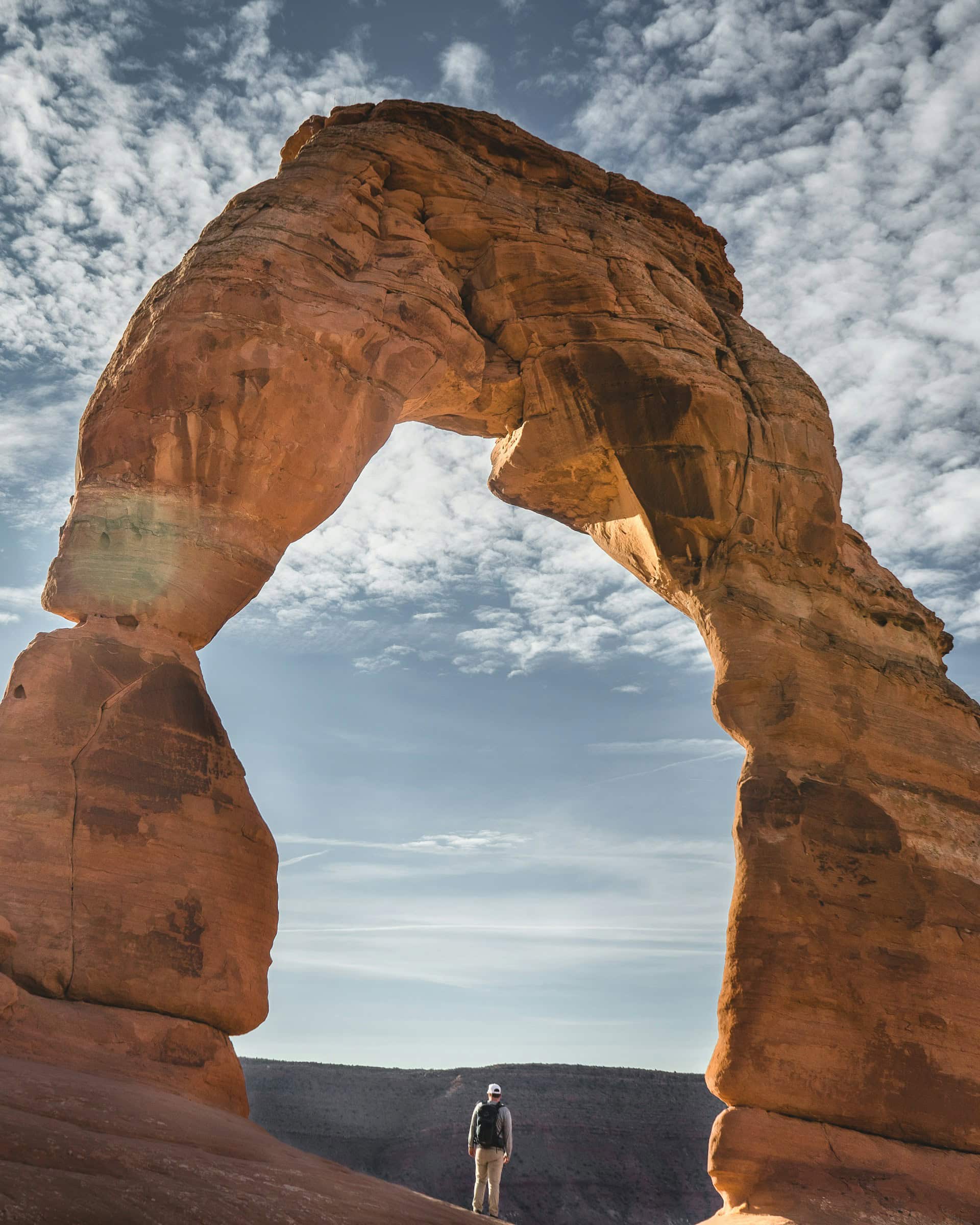 A large stone arch against a blue sky with clouds. A person in a hat and backpack facing away stands in the middle underneath the arch.