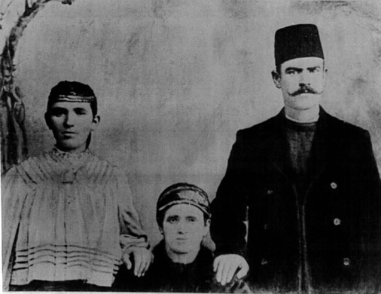 A copy of a black and white photograph of a man and a woman standing, each with a hand on the shoulder of a third, presumably seated figure (the image cuts off at their chest).