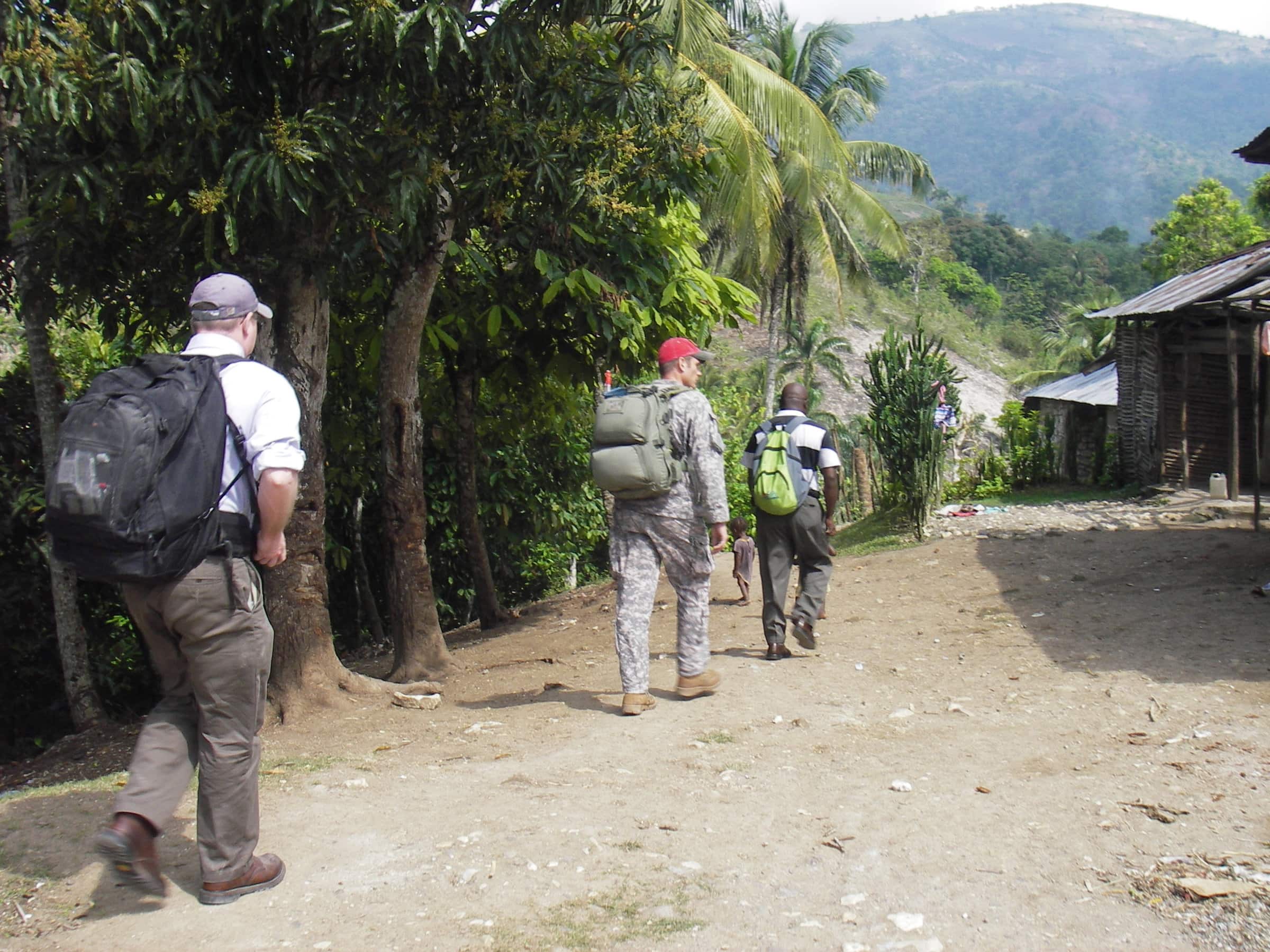 Troy Sacquety in grey pants, a white button down shirt, a navy baseball cap, and black backpack accompanies a Civil Affairs Team member in an army uniform and red baseball cap. They are walking on a dirt road with wooden houses on the right and tropical trees on the left.