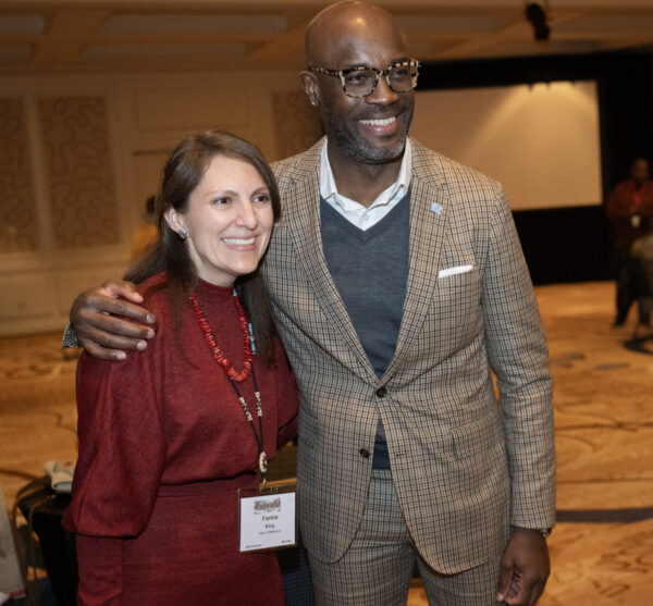 A white woman wearing a red dress next to a Black man wearing a tan suit embracing and smiling for a picture
