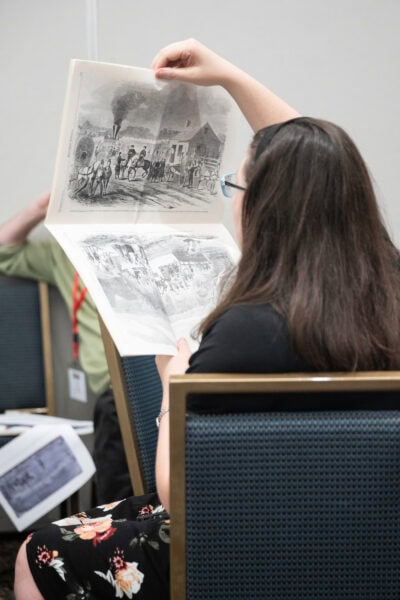 The back of a woman sitting and holding a newspaper with an illustration close to her face