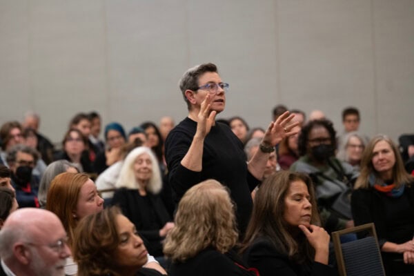 A woman with short hair and glasses standing up and speaking in the middle of a seated audience