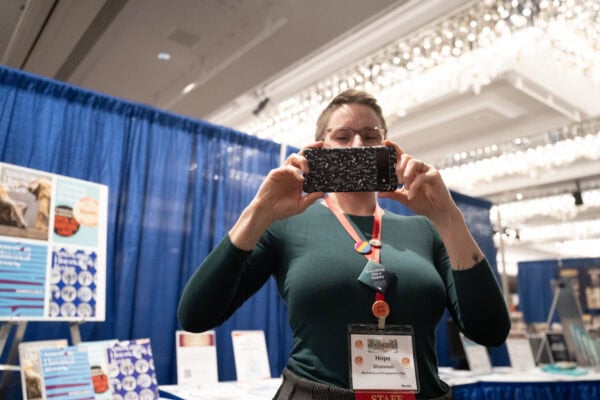 A white woman wearing a green top holding an iPhone over her face pointed towards the camera