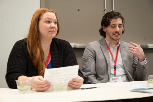 A white woman with long red hair holding a paper next to a white man speaking on a panel