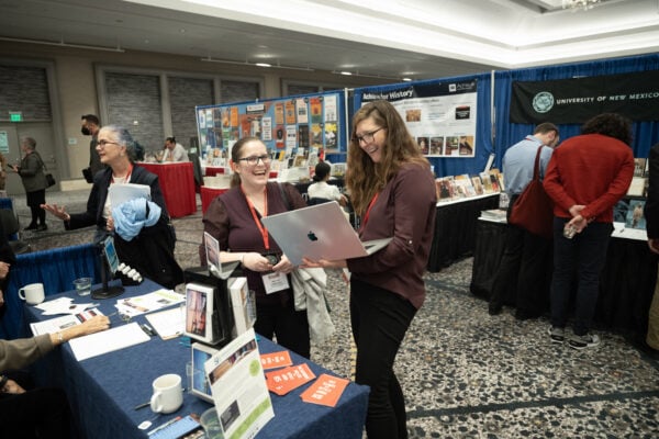 Two women standing at a booth laughing. The one on the right is holding a laptop