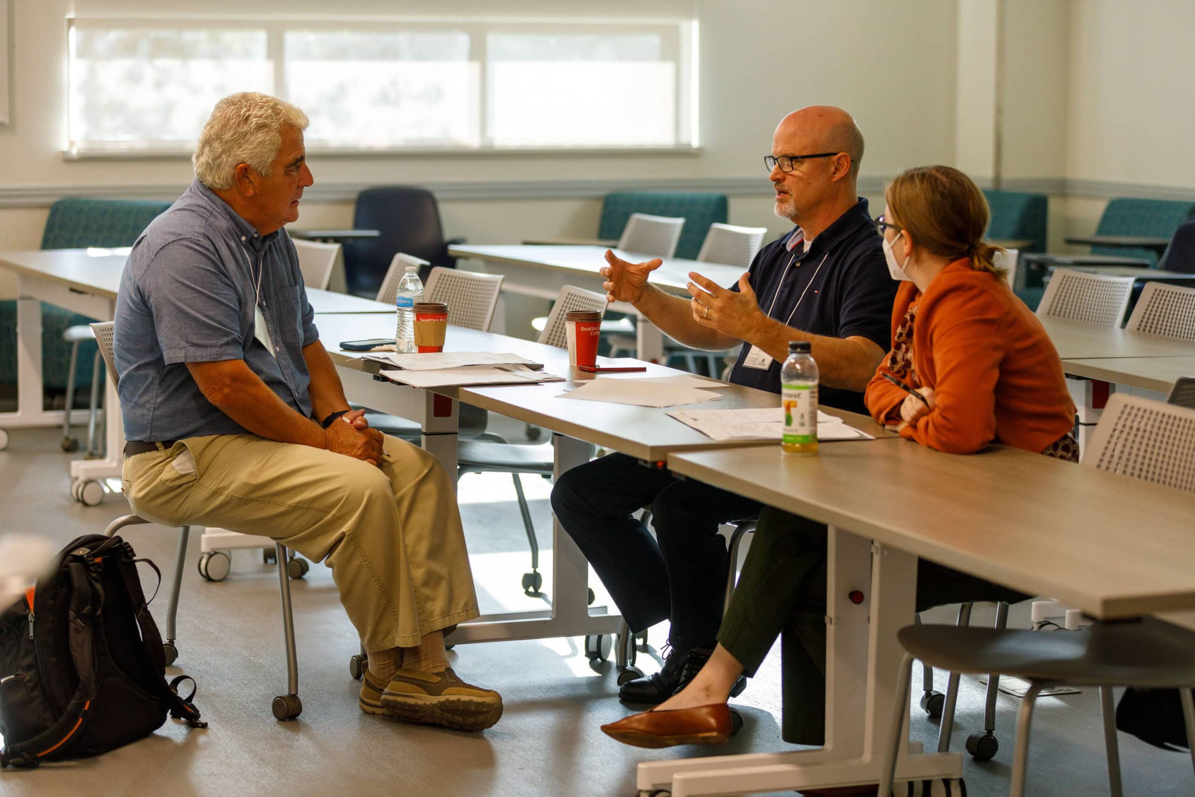 Two men and one woman are sitting around a table in a discussion group. There are papers and coffee cups in front of them.