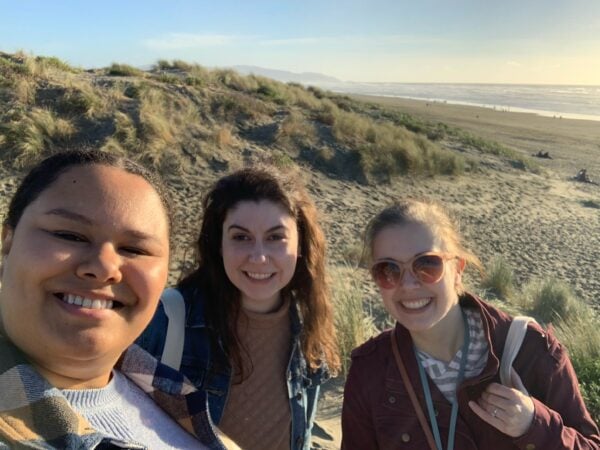A selfie of three women smiling with a beach behind them