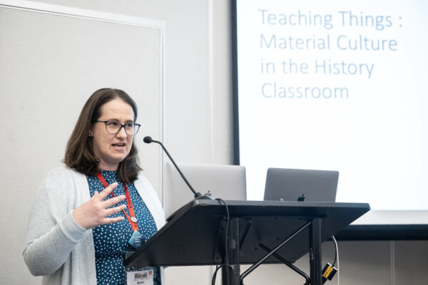 A white woman with glasses speaking at a podium with a powerpoint behind her